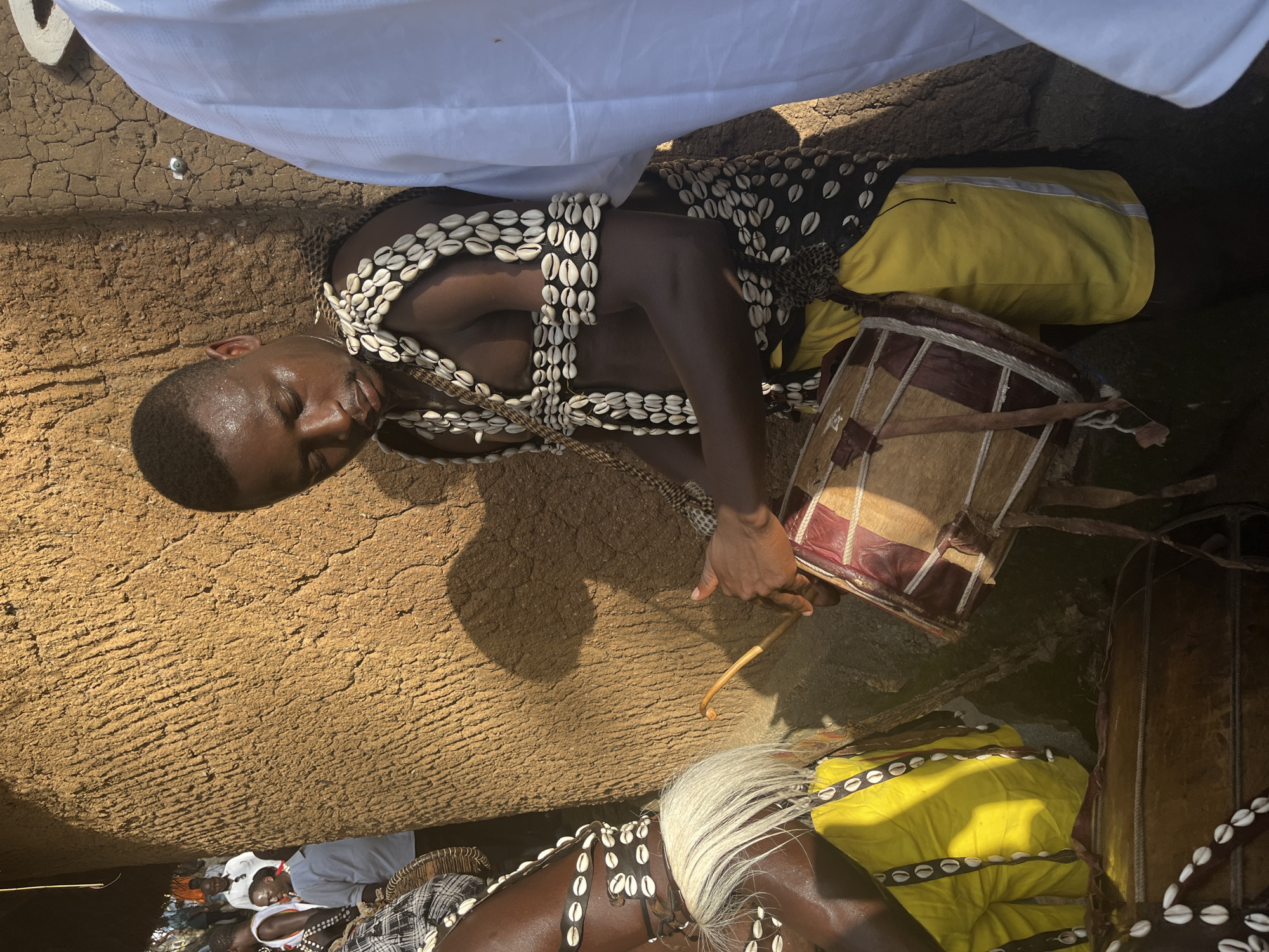 The image depicts a scene in Jos, Nigeria, possibly during a cultural or spiritual gathering. The central subject is a person adorned with multiple necklaces of white shells. They are lying down with their eyes closed, appearing to be in a state of deep concentration or trance. A drum, with a natural hide head and a wooden frame, is placed beside them, and the person's hand is positioned as if holding a drumstick, suggesting they might have been or are about to play it.
Another individual, also wearing shell adornments and a yellow garment, is partially visible in the lower part of the frame, with their back to the camera. This suggests a communal aspect to the event. The ground appears to be dry, cracked earth, indicative of a warm, possibly arid climate, and the lighting suggests it's daytime, with sunlight casting shadows. The overall atmosphere seems solemn and ritualistic. There is no visible text in the image.