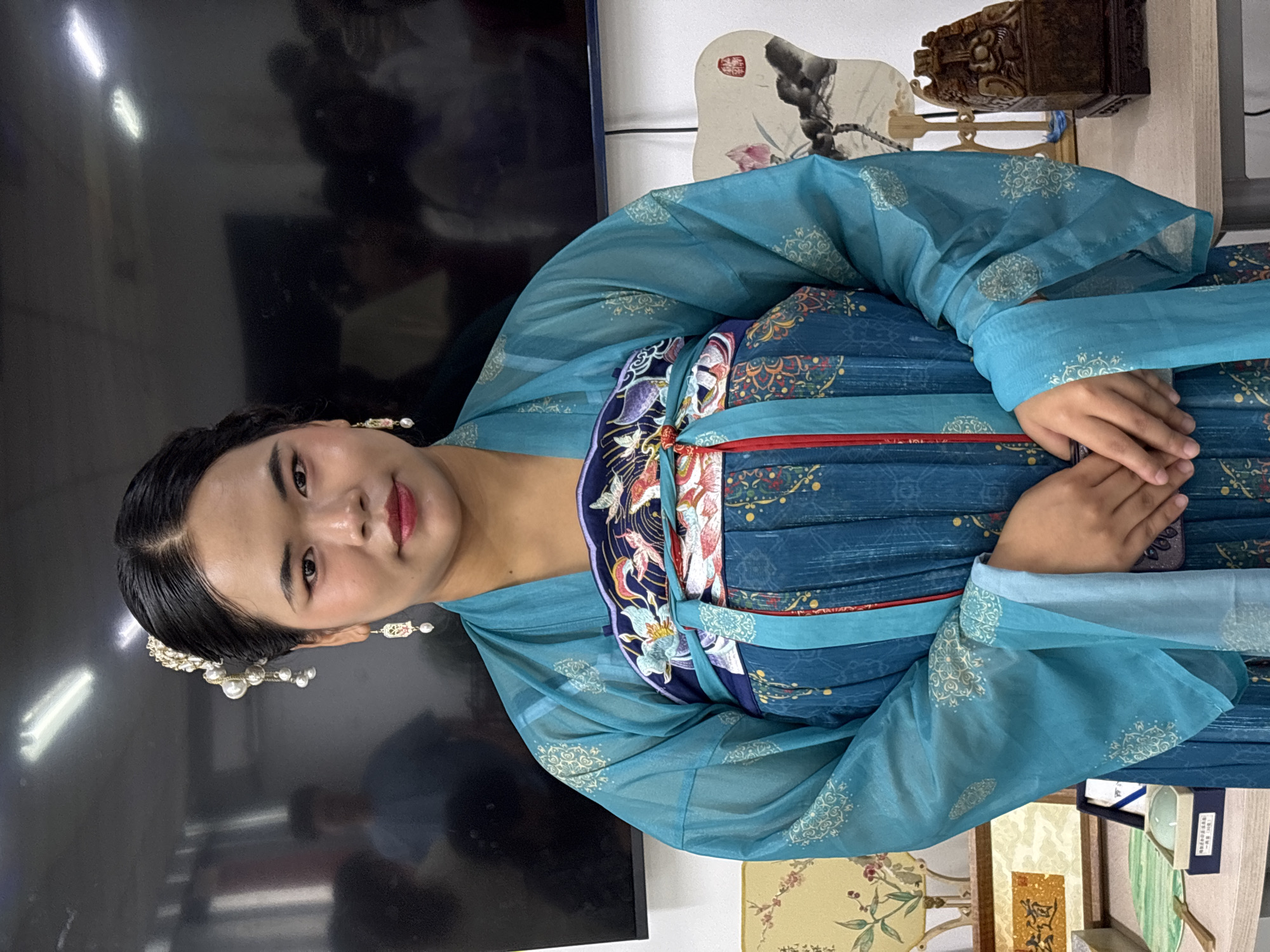 A woman wearing a traditional blue Chinese dress and hair ornaments poses indoors. She is seated or standing against a dark background that includes a television screen and various decorative items like a fan, an ornate wooden box, and a framed display. The setting appears to be in a domestic or studio environment. It is not possible to determine whether or not the location is in Jiangjin, China.