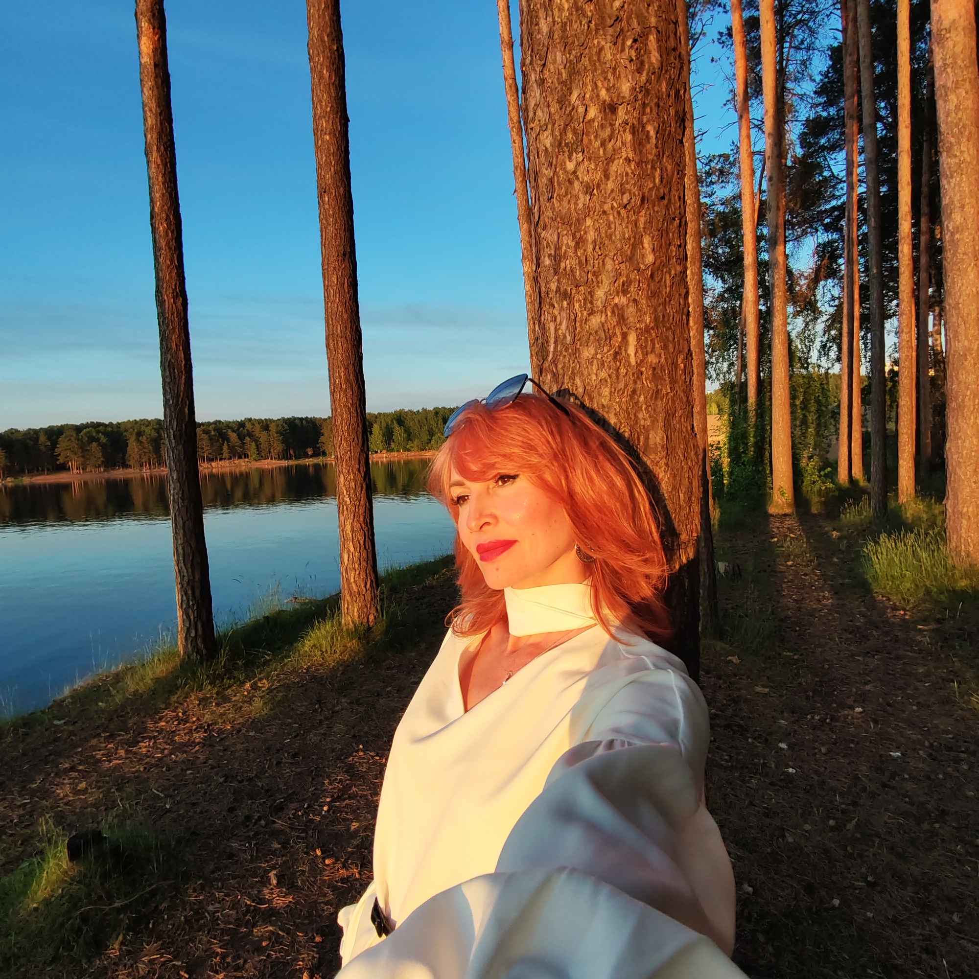 This media file depicts a woman in a serene natural setting in Chaikovsky, Russia, during what appears to be golden hour. The main subject is a woman with reddish-orange hair, seen in a close-up, leaning against a large pine tree trunk. She is wearing a light-colored (possibly white or cream) long-sleeved top with a high collar or scarf, and sunglasses are perched on top of her head. Her face is illuminated by warm, golden sunlight, highlighting her features and pink lipstick, and she has a calm, contemplative expression, looking off to her left.
The setting is a pine forest adjacent to a tranquil body of water, likely a lake or wide river. The water reflects the clear blue sky and a distant treeline, suggesting a calm surface. Several other pine trees with reddish-brown bark are visible in the foreground and background, their trunks catching the warm light of the low sun. The sunlight casts long shadows across the forest floor, which is covered with pine needles, grass, and some small pinecones. The sky above is a vibrant, clear blue. The overall atmosphere is peaceful and picturesque, with the warm light creating a dramatic and inviting scene. The woman appears to be posing for a photograph, possibly a selfie. There is no visible text in the image.