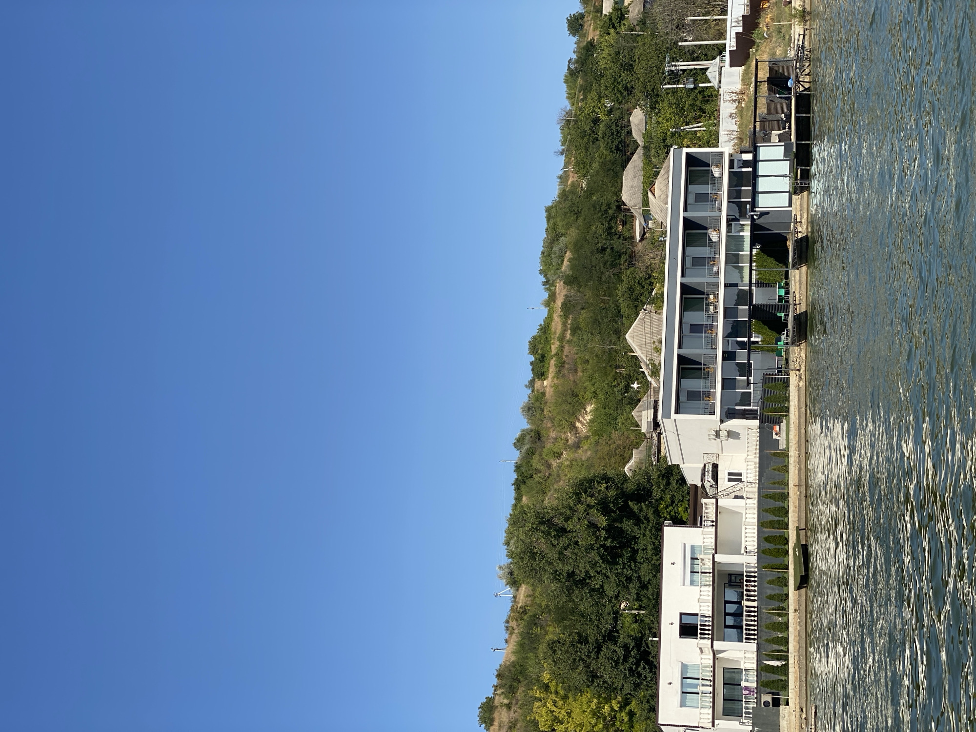 A wide-angle, eye-level, outdoor shot captures a waterfront property on a sunny day with a clear blue sky. In the foreground, rippling greenish-blue water occupies the lower third of the frame. The middle ground features a modern, multi-story building with white and grey facades, situated directly on the water's edge. This building has multiple balconies with railings and large glass doors. Adjacent to it, there is a lower section of the building with a darker facade, featuring a covered outdoor area with tables and chairs. A wooden deck with a ladder extends into the water. Several small, flat-bottomed boats are moored near the deck. A small, green boat rests on a submerged wooden structure in the water to the left of the main buildings. A neatly trimmed row of green conical bushes lines the edge of the property between the building and the water. The background showcases a steep, tree-covered hillside with a few scattered houses and a dirt path leading up. Electrical wires and poles are visible on the hillside. The scene appears to be the shore of the Nistru River in Molovata, Moldova. No people are visible in the image.