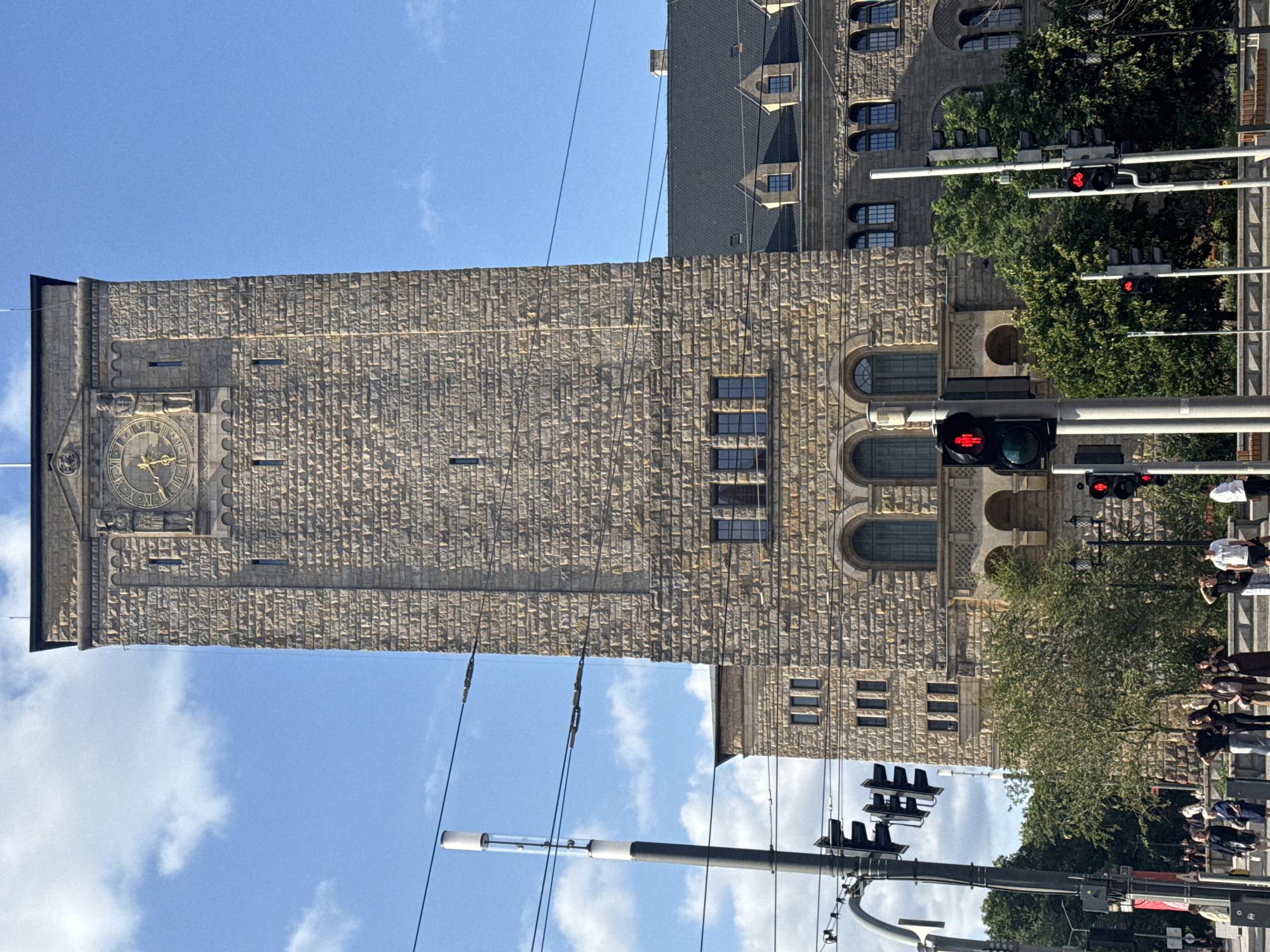 This is a daytime, eye-level, full-shot photograph of the Poznań City Hall tower in Poznań, Poland. The imposing tower, constructed of light brown stone blocks, dominates the frame. The tower features a large clock face near its apex, adorned with Roman numerals and flanked by two sculpted figures. Below the clock, there are several arched windows and decorative architectural elements. The building is surrounded by trees and a clear blue sky with some clouds. In the foreground, there are traffic lights and a street scene with pedestrians. The overall impression is one of grandeur and historical significance within a bustling urban environment.
