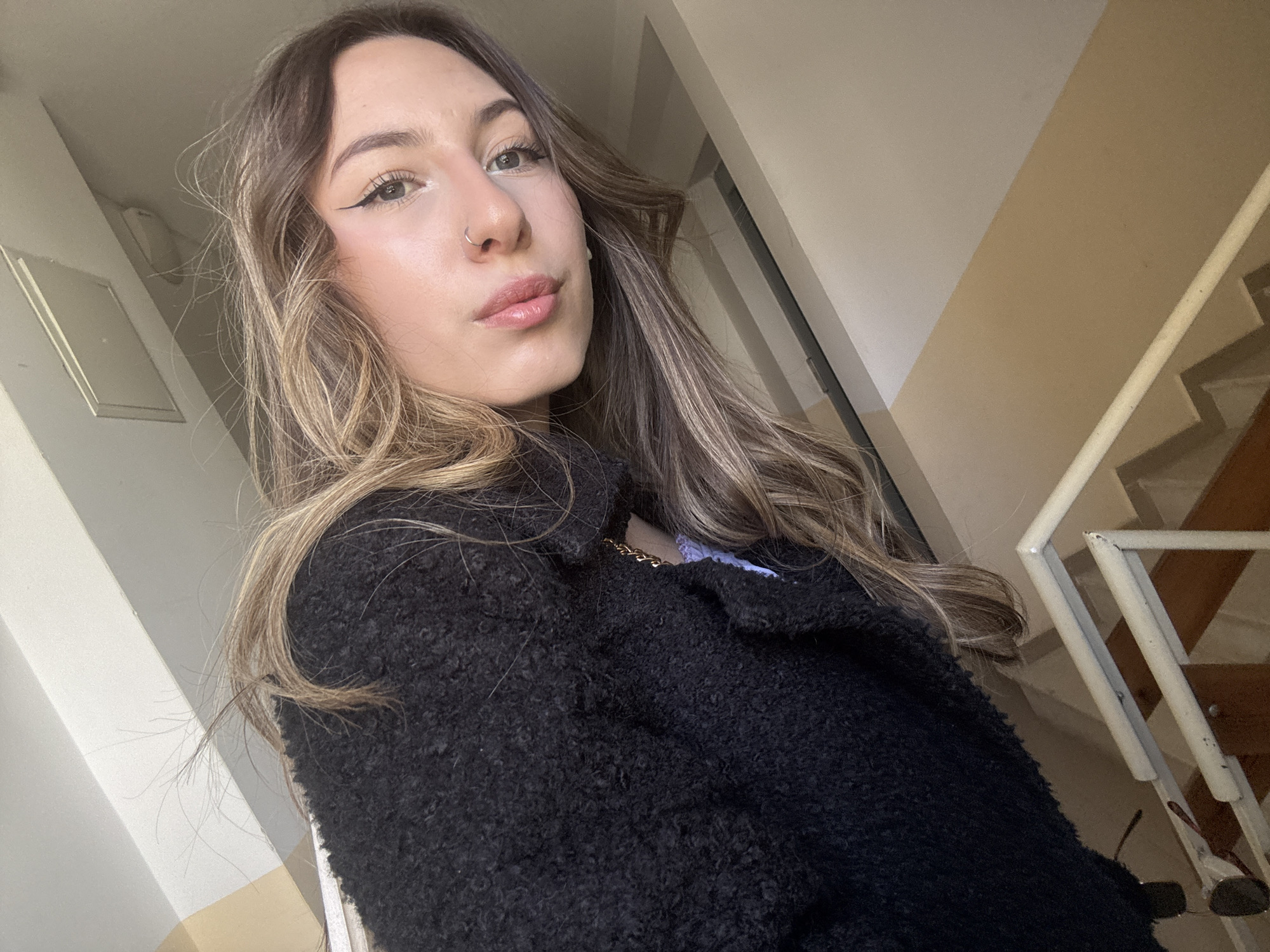 A young woman is featured in a close-up, angled portrait, likely a selfie, taken indoors in Veliko Tarnovo, Bulgaria. She has long, wavy brown hair with lighter highlights, framing her face. Her makeup includes precise winged eyeliner and a subtle gloss on her full lips. A small silver hoop nose ring is visible on her left nostril. Her expression is neutral as she looks towards the camera.
She is wearing a textured black jacket or coat, possibly made of boucle or fleece-like material, with a hint of a light purple garment visible underneath near her neck, complemented by a thin gold chain.
The background suggests an interior setting, likely a hallway or stairwell. To her left, there's a light-colored wall with a small rectangular panel or switch. To her right and behind her, the background consists of a two-toned wall (beige and off-white) and a partial view of a white metal railing and wooden stairs, indicating a staircase. A pair of sunglasses can be seen hanging on the railing in the lower right corner. The scene is lit by bright natural light, suggesting it is daytime. There is no visible text in the image.