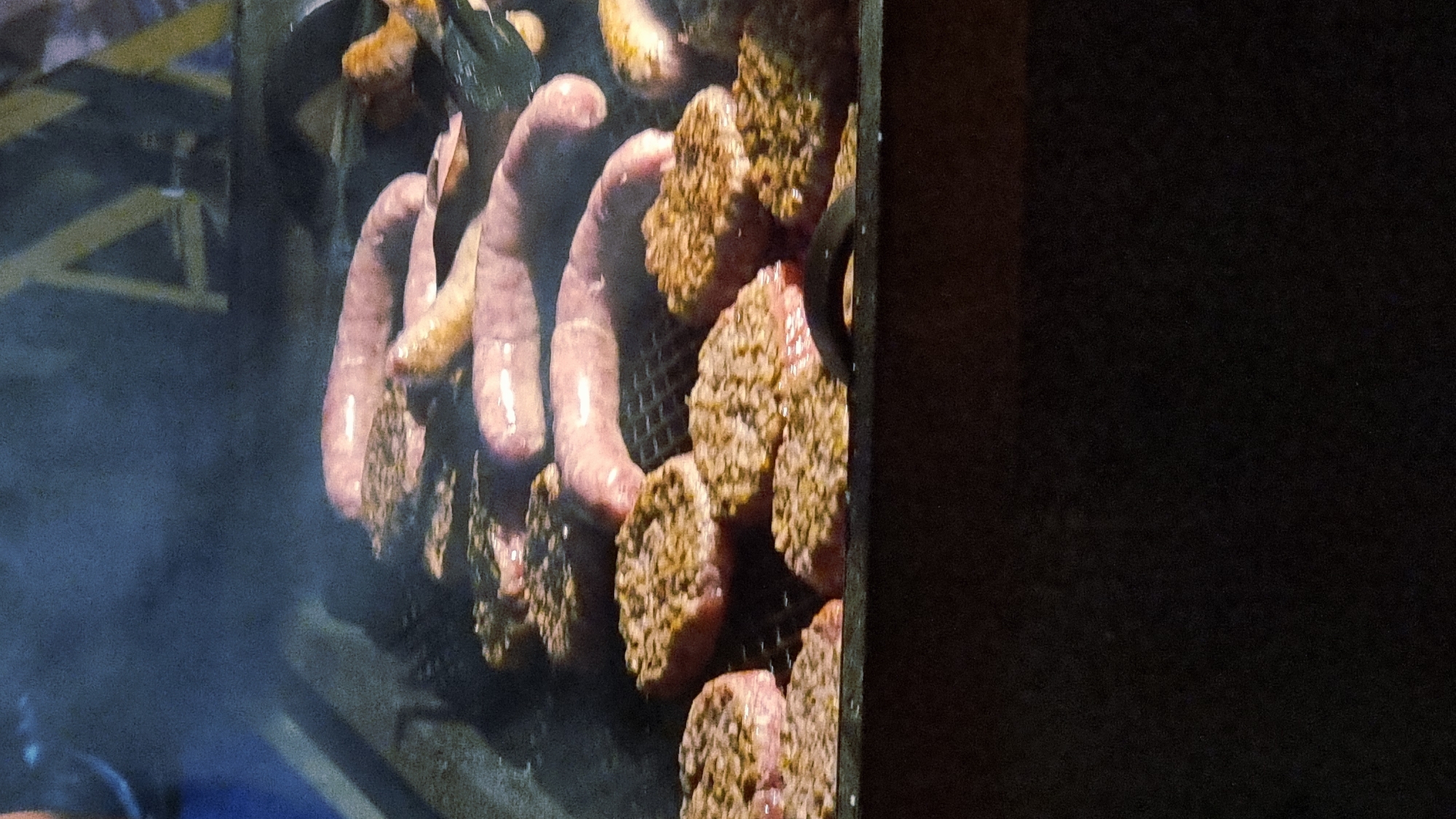 The image depicts a close-up view of a grill laden with food, including several uncooked sausages and numerous ground meat patties. The sausages are a pale pink color and appear to be linked together, some curved and overlapping. The patties are round and flat, with a textured surface suggesting they have been seasoned. Wisps of smoke rise from the grill, indicating the food is actively cooking. The grill itself is made of metal grates, and a portion of its dark, textured side is visible at the bottom of the frame. The background is out of focus, showing blurred shapes of yellow structures that might be part of an outdoor setup. No people are directly visible in the frame. The overall impression is that of food being prepared on a barbecue. The context of Sant Joan Les Fonts, Spain is not discernible from the visual elements of the image itself.