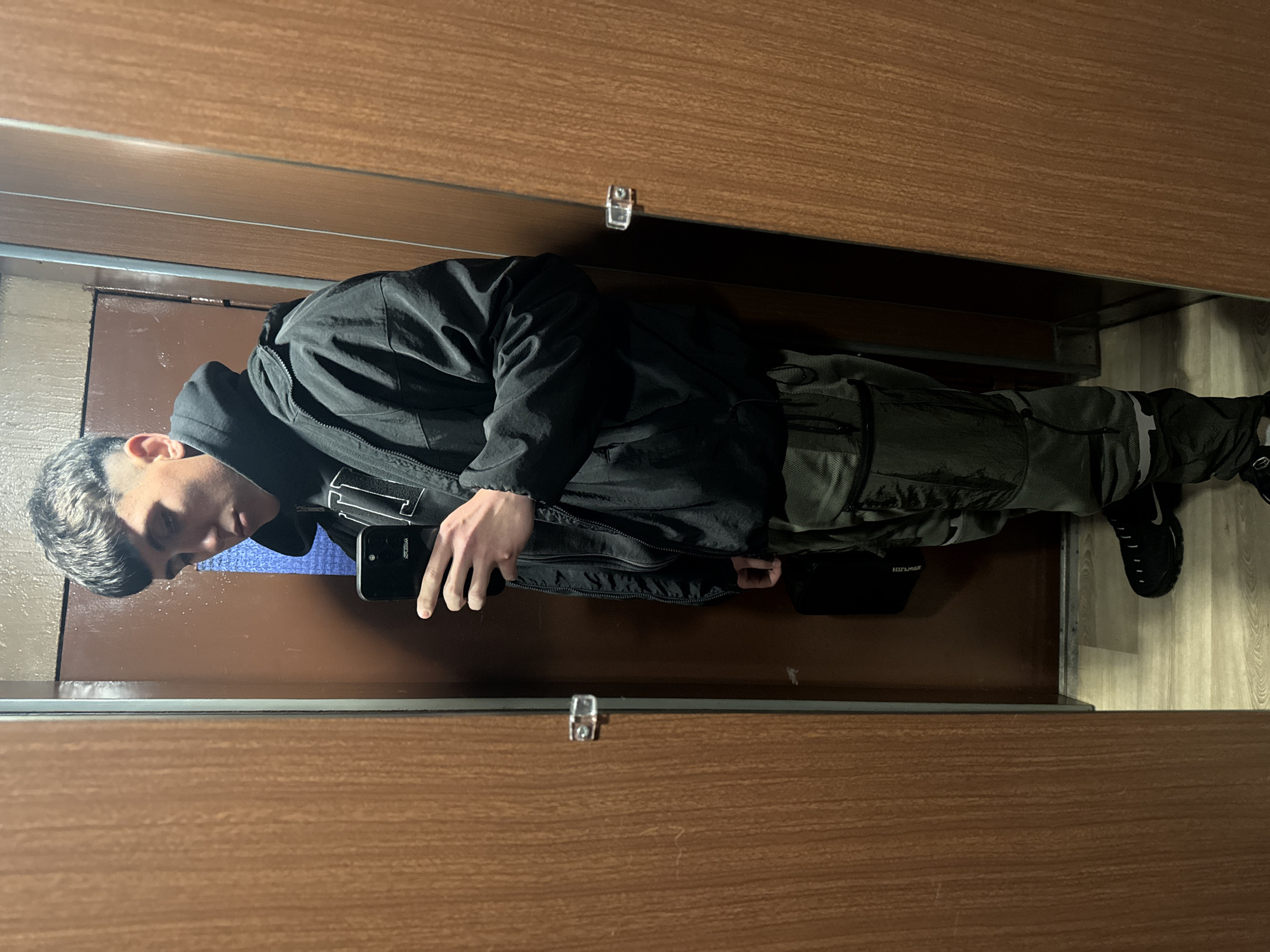 A young man is taking a selfie in what appears to be a small elevator. The walls of the elevator are made of wood panels. The man is wearing a black hooded jacket, dark-colored pants with lighter gray accents, and black sneakers. He is holding a black smartphone in his right hand to take the photo. The elevator floor is visible, showing a light-colored wooden surface. The location of the photo, Varna, Bulgaria, cannot be independently determined based on the visual information provided.