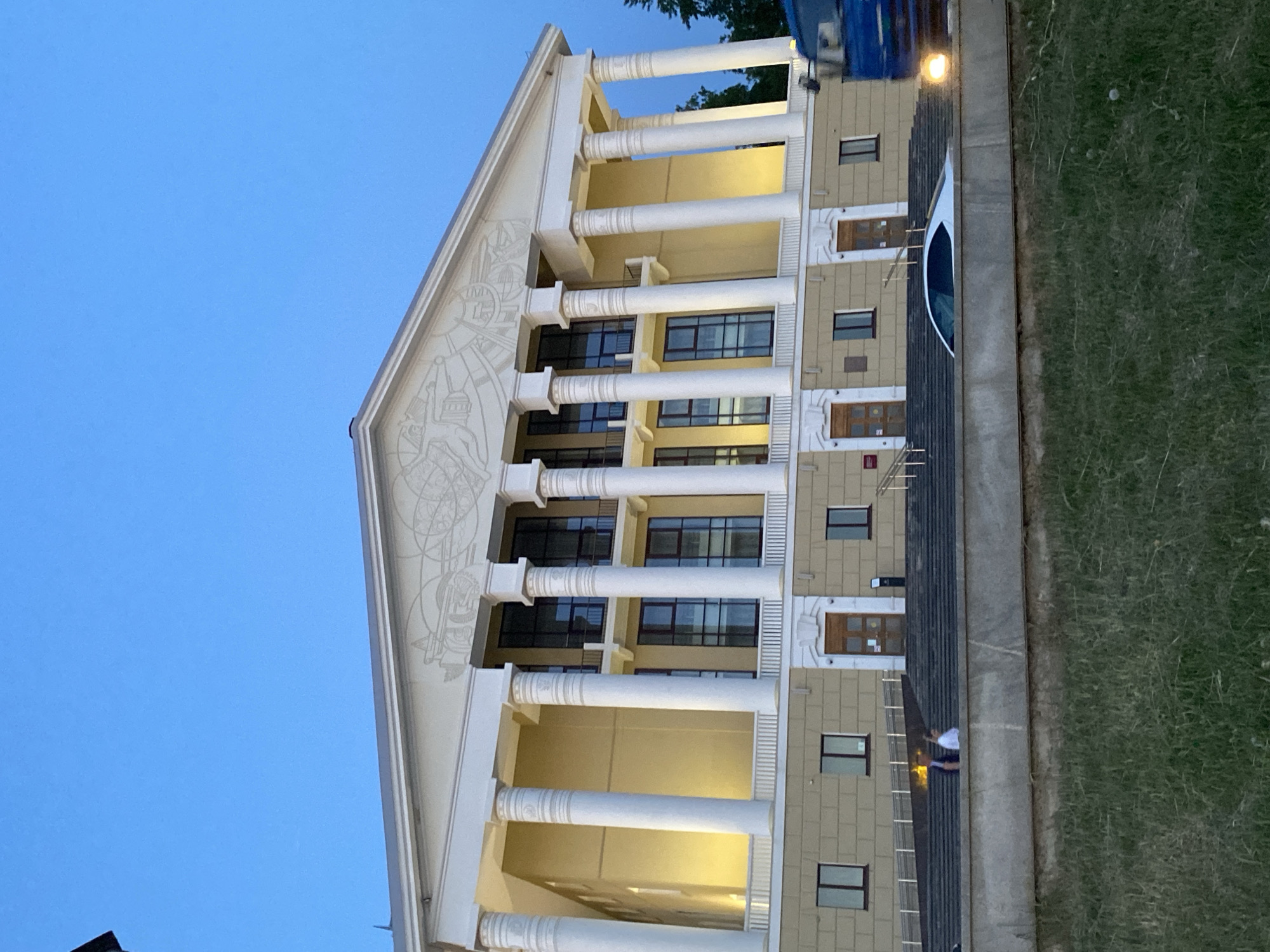 The image shows the exterior of the House of Culture building in Votkinsk, Russia, during evening twilight. The building has a classical architectural style, featuring a pediment with decorative relief, a colonnade of white columns, windows, and exterior lighting. A parked van or bus is visible near the building, with a section of sidewalk, a metal railing, and grassy area adjacent. A person is walking on the steps adjacent to the building. The sky is a clear, deep blue.