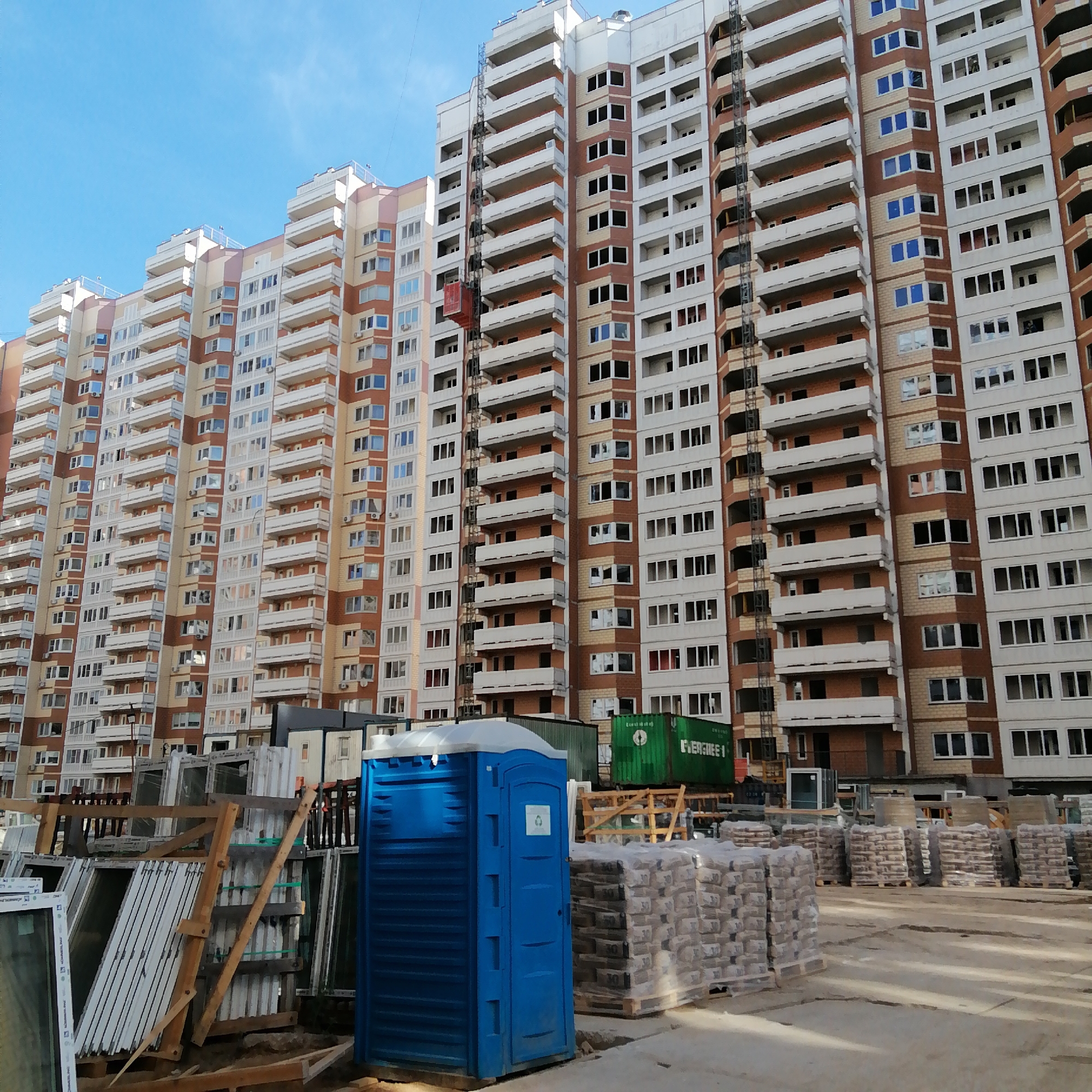 The image shows a construction site in Unknown, Unknown with a tall, multi-story building in the background. The building is predominantly white with brick-colored accents. A construction elevator is visible on one side of the building.
In the foreground, there are construction materials, including stacks of bagged goods, windows, and a blue portable toilet. A green Evergreen shipping container is partially visible behind the construction materials. The ground appears to be a paved construction area.