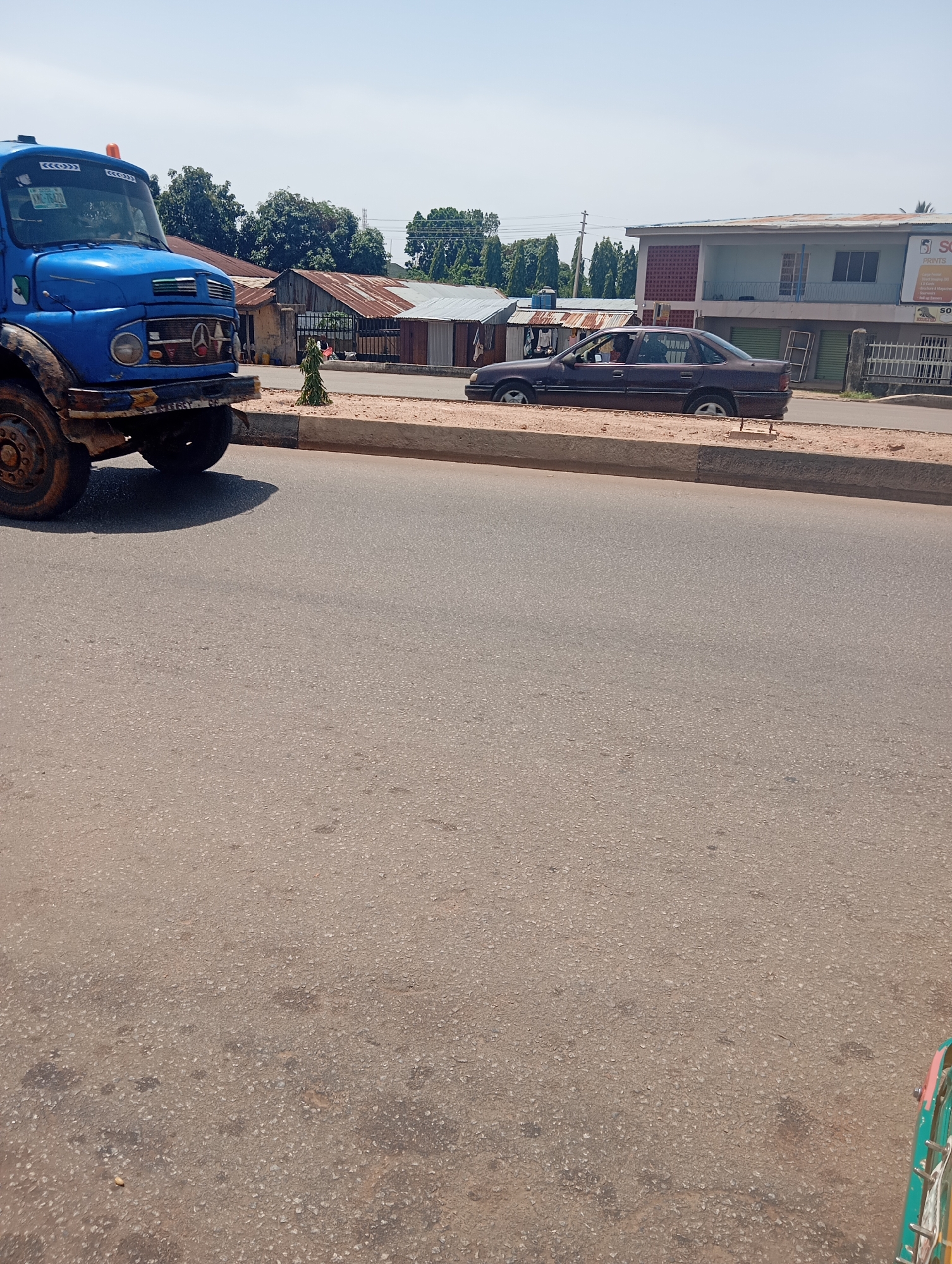 This media file shows a street scene in Jos, Nigeria, on a bright, sunny day. The foreground is dominated by the asphalt road, showing some wear and tear with patches of darker material. On the left side of the image, a bright blue truck is partially visible, with its large tire and mudguards prominent. The truck appears to be in motion or has recently stopped.
Further down the street, a dark-colored sedan is parked or slowly moving. The sedan has a tinted windshield and the occupants are not clearly visible. Behind the vehicles, a row of buildings lines the street. These buildings vary in style, with some featuring corrugated metal roofs and others appearing to be more permanent structures with painted facades. One building, towards the right, has a sign that is partially visible, with the letters "SC" and "SON" being discernible. This suggests a commercial area.
Trees with lush green foliage are scattered throughout the background, indicating a well-vegetated environment. The sky is a clear, pale blue, reinforcing the impression of a sunny day. The overall atmosphere appears to be one of everyday activity in a bustling urban or semi-urban setting in Nigeria. No specific events are depicted, but the presence of vehicles and buildings suggests normal daily operations.