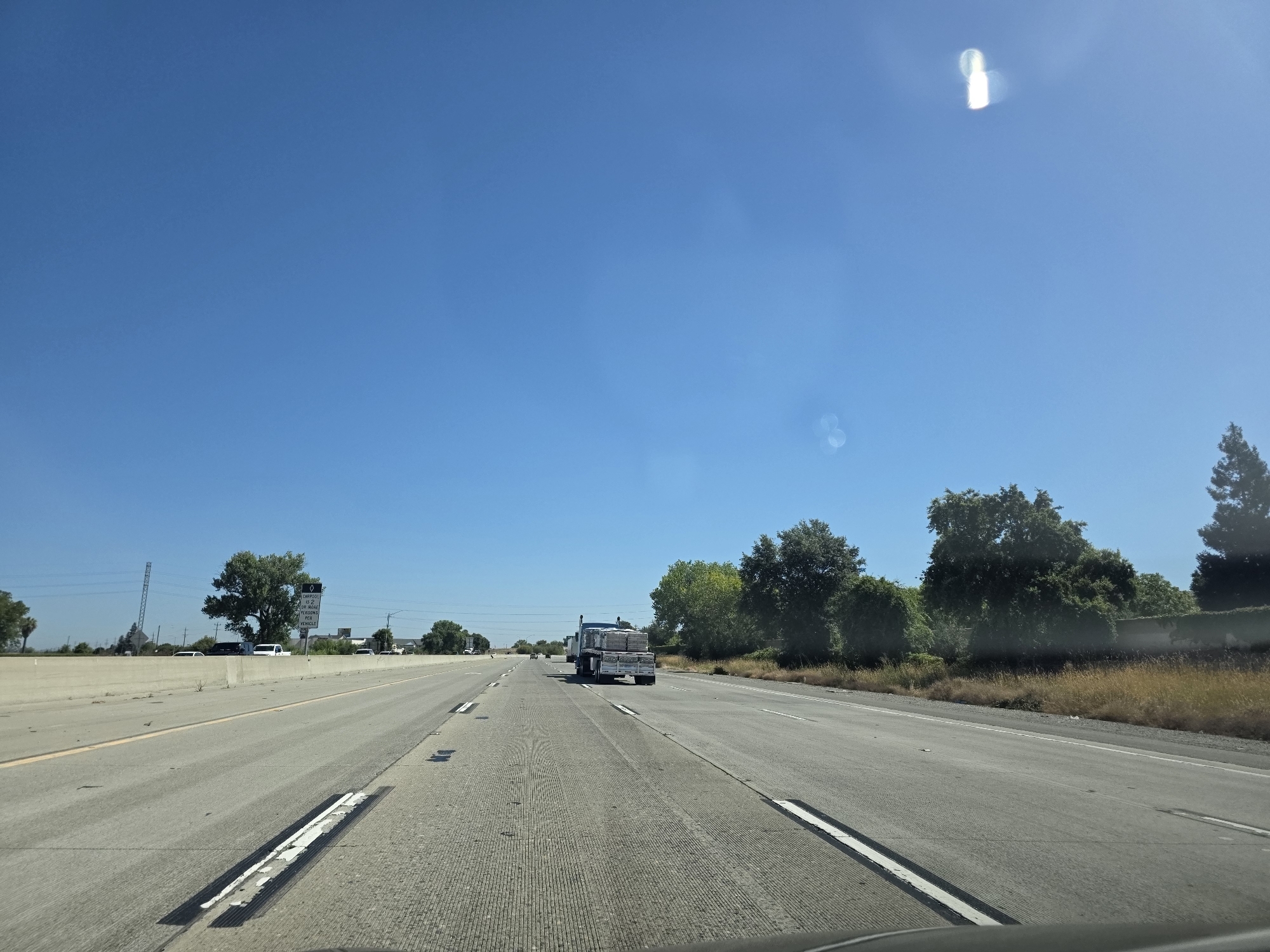 The media file shows a highway scene in Sacramento, California, under a clear blue sky, indicating daytime. The perspective is from inside a vehicle, suggesting the shot was taken while driving. The road is paved, with multiple lanes visible. On the left side of the road, there's a concrete barrier and beyond that, some greenery and power lines.
Further down the road on the left, a white sign is visible. Zooming in on the sign reveals text that appears to be regulatory, possibly related to vehicle restrictions or speed limits, although the details are difficult to discern. A truck is seen in the right lane of the highway ahead, carrying a load of what appear to be stacked materials, possibly building supplies. The truck is a semi-trailer truck.
To the right of the highway, there is a grassy area with dry brush, bordered by trees and a low wall. The overall impression is a typical highway commute in a semi-arid or warm climate, given the dry vegetation and clear skies. There are no people clearly visible in the frame, nor any explicit indication of a specific event. The scene conveys a sense of travel and the open road. There are some lens flares visible in the sky, indicating the sun is bright.