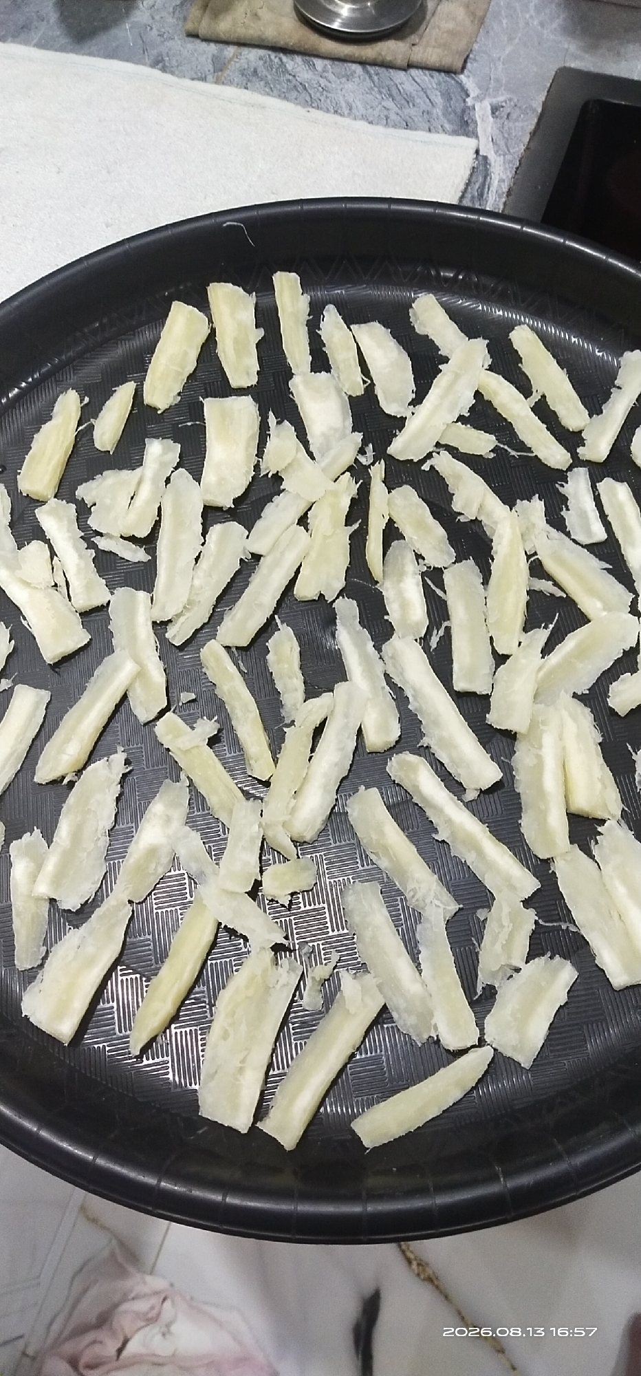 A close-up, overhead shot captures a black, textured tray filled with numerous strips of peeled, white cassava. The cassava pieces are cut into irregular, elongated shapes, some appearing more fibrous and rough than others. They are arranged somewhat haphazardly across the entire surface of the tray, with some overlapping. The background is blurred, showing a countertop and a white cloth. A timestamp in the lower right corner reads "2026.08.13 16:57". The location is identified as Matabu, Indonesia.