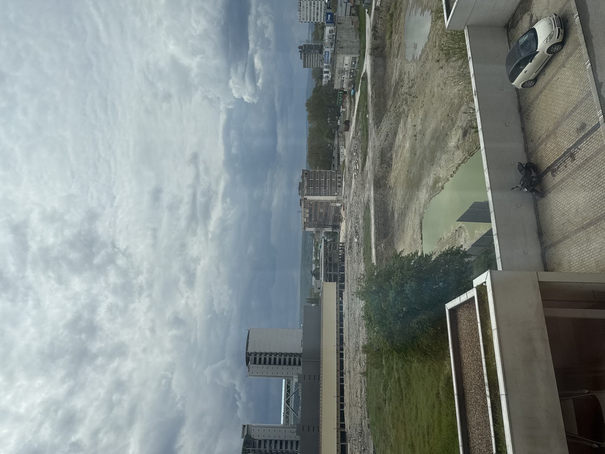 The image, taken from an elevated perspective and oriented vertically, captures a sprawling urban landscape under an overcast sky. The top half of the frame is dominated by a vast expanse of light grey, cloudy sky, suggesting a heavily clouded or pre-storm weather condition during daytime.
In the middle ground, a mix of developed and undeveloped areas is visible. Several multi-story buildings, appearing to be residential or commercial, dot the horizon. One building clearly displays the name "DECATLON" (Decathlon), indicating a retail store. Another building shows partially visible text, possibly "TYCK". Beyond the immediate buildings, a wide swath of barren, disturbed earth, likely a construction site or recently cleared land, stretches across the mid-section. Within this area, a distinct pool of stagnant, green-tinged water is noticeable. Scattered patches of green grass and a few trees break up the dry earth. In the far distance, faint outlines of hills or mountains are visible under the cloudy horizon.
The foreground of the image shows architectural elements of the building from which the photograph was taken, including a concrete ledge or balcony and a planter box filled with dark, granular material. Adjacent to this structure, on a paved surface with block patterns, a white compact car (resembling a Smart car) is parked. Nearby, a black motorcycle is also parked on the paved ground, where parking space markings like "L6" and "L5" are visible. No human activity or individuals are discernible in the scene, which presents a static overview of an urban environment, likely in Varna, Bulgaria, characterized by ongoing development or land preparation.