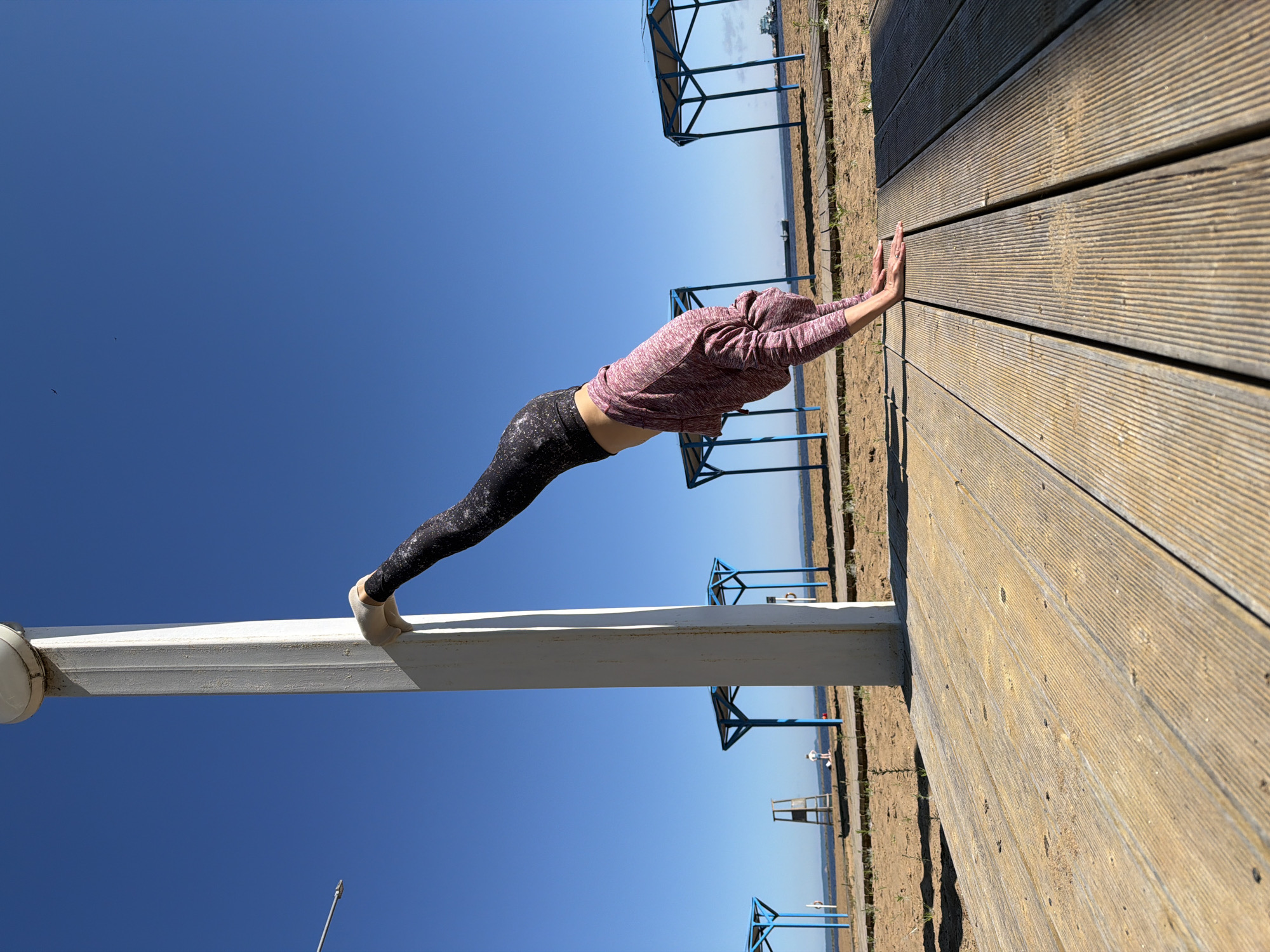 The image depicts a woman performing a yoga pose, specifically a variation of downward-facing dog, with her feet elevated and resting against a white pole. She is on a wooden walkway at a beach, with sand and grass visible. In the background, there are several blue and white structures, likely beach shelters, and a body of water that appears to be a reservoir or river. A few people are scattered in the distance, and two birds are flying in the clear blue sky. The overall scene is set in a bright, sunny outdoor environment. The location is identified as Chaikovsky, Russia.