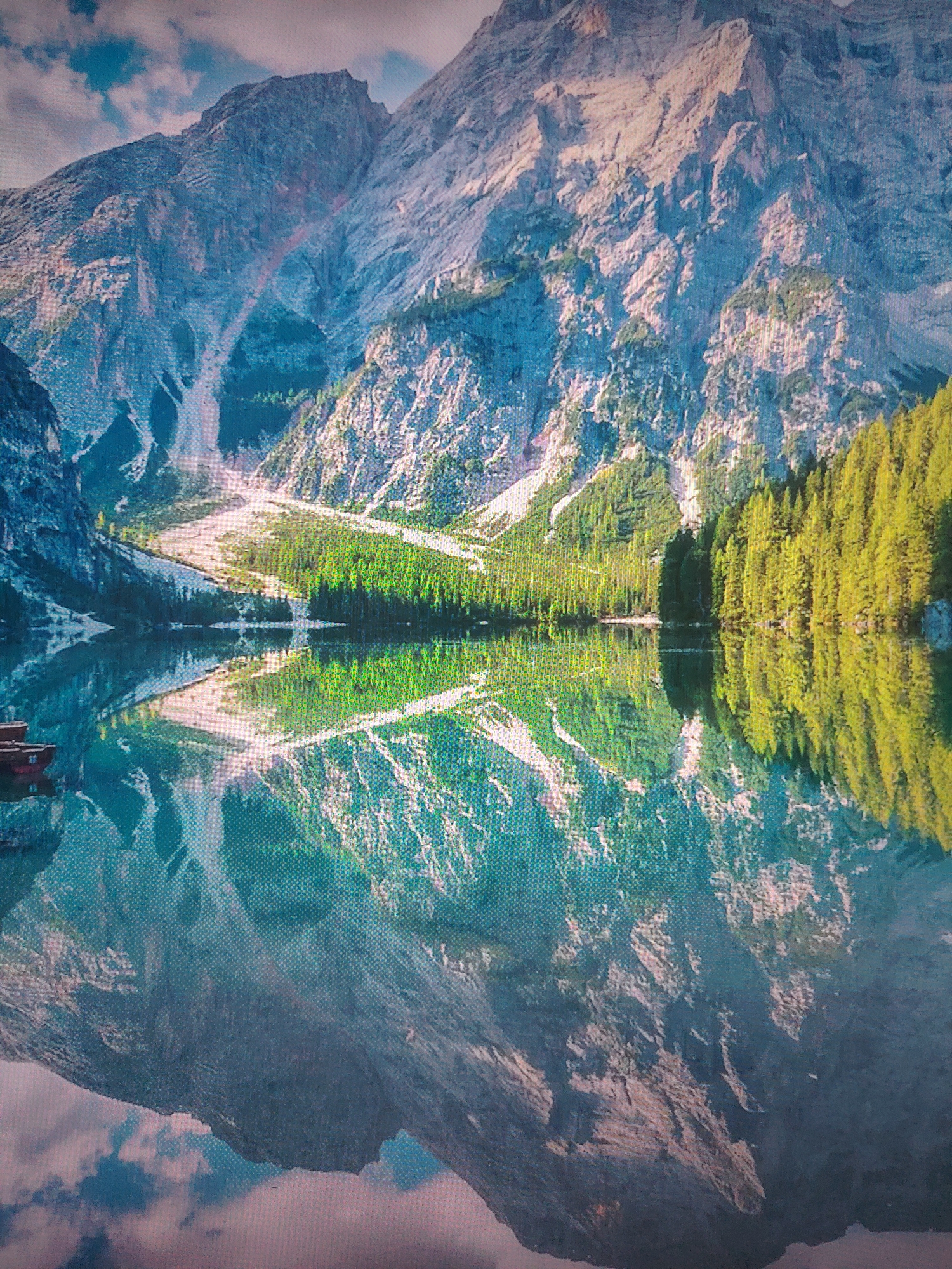 The image shows a mountain reflected in a lake, surrounded by trees. The sky is partly cloudy. There are several boats visible in the left side of the lake. Location is in the city of Unknown, Unknown.