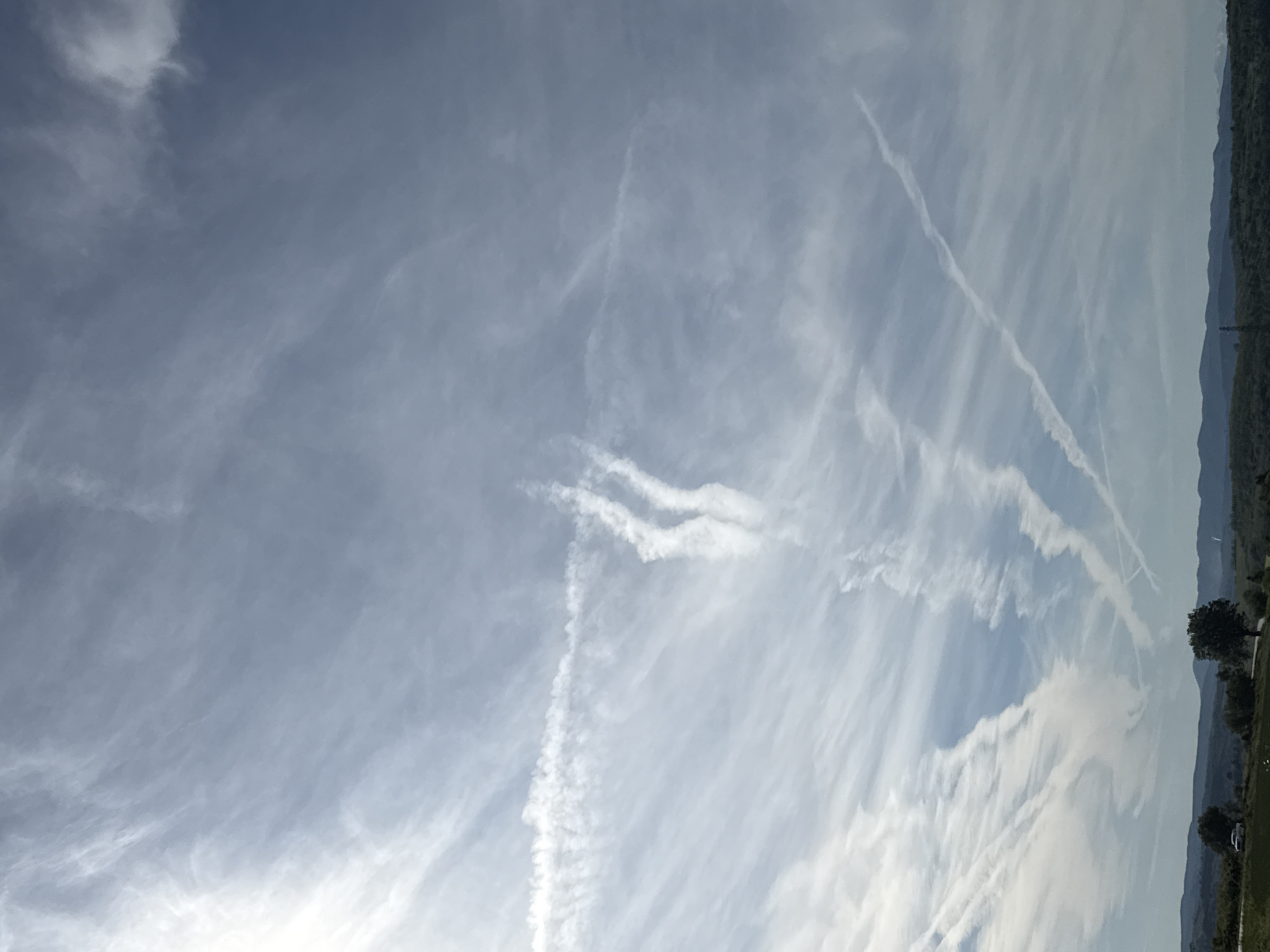 The image shows a daytime sky with numerous contrails. The sky is pale blue with wispy cirrus clouds and contrails that intersect and dissipate. The horizon line is visible in the upper right corner, suggesting a distant landmass or hills with sparse vegetation. The location is Ugarchin, Bulgaria.