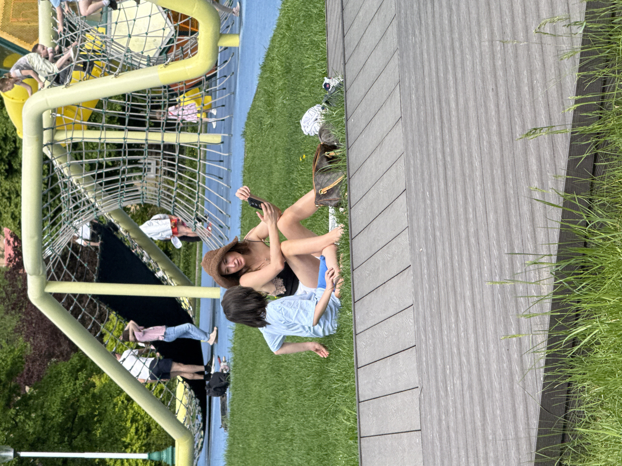 The image depicts a scene at Zaryadye Park in Moscow, Russia. A woman wearing a brown hat and a black and white top is sitting on the grass, looking at a phone she is holding. A child, presumably a boy, wearing a light blue t-shirt is sitting next to her and appears to be interacting with her. A designer handbag is near them on the grass. Behind them is a yellow playground structure with a net. Other people are visible in the background, some walking and others on the playground equipment. A grey wooden deck with grass growing along its edge is visible on the right side of the image. The overall environment seems to be a sunny outdoor recreational area.