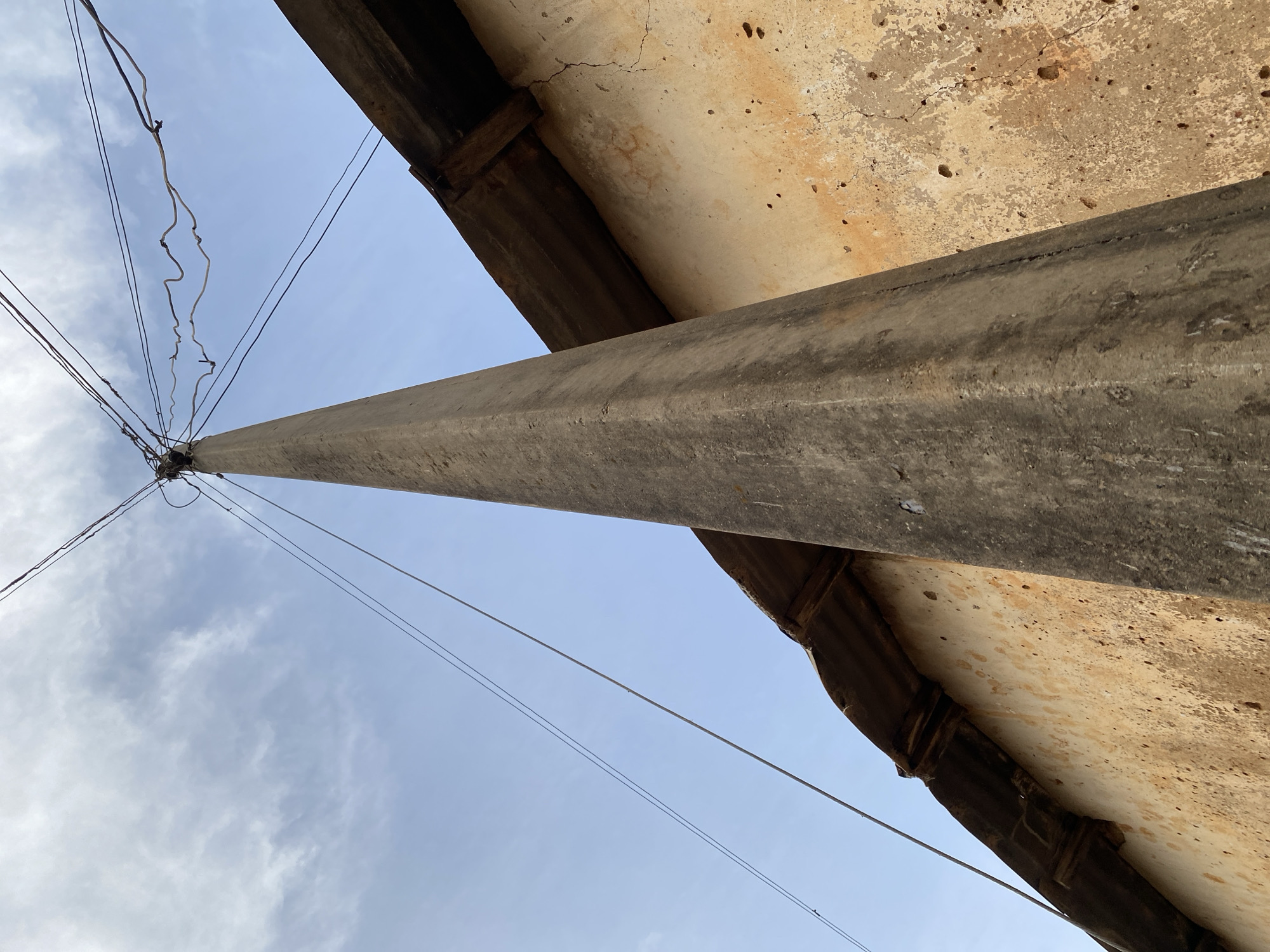 The image is an upward-facing shot of a concrete utility pole against a sky with scattered clouds. The pole is thick and extends diagonally across the frame, appearing to be made of rough, weathered concrete. Several electrical wires are strung from the top of the pole, creating a tangled network against the sky.
To the right of the pole, a textured, aged building facade is visible. This wall appears to be made of stucco or a similar material, showing signs of wear with cracks, discoloration, and small holes. A darker, ribbed structure, possibly a gutter or roof edge, runs along the bottom of this facade.
The overall impression is one of an outdoor, possibly urban or semi-rural setting, given the utility pole and building. The lighting suggests it is daytime, and the sky's appearance indicates fair weather. There are no people or specific events depicted. The focus is on the structural elements of the utility pole and the adjacent building, contrasted with the open sky. No text is visible in the image. The image appears to have been taken in Bukuru, Nigeria.