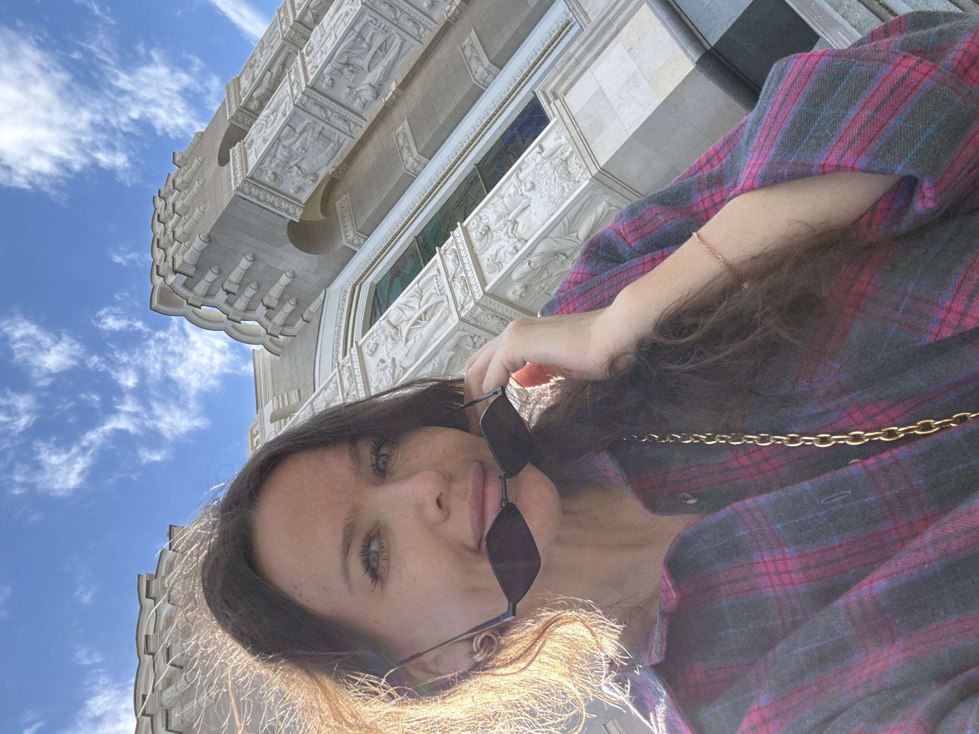 The media file shows a low-angle selfie of a young woman with long, dark hair and striking blue eyes, taken outdoors. She is wearing a dark plaid shirt with pink and blue lines, and a gold chain necklace. She is holding a pair of dark-rimmed sunglasses in her right hand. Her expression is pleasant and she appears to be smiling.
The background features an ornate, pale stone building with intricate carvings and architectural details, possibly a temple or a historical monument. The sky above is a vibrant blue, dotted with scattered white clouds, suggesting it is daytime with good weather. The sunlight is casting a warm glow on the woman's hair. The image has a slightly tilted perspective, emphasizing the grandeur of the building. The scene is set in Moscow, Russia. There is no visible text in the media file.