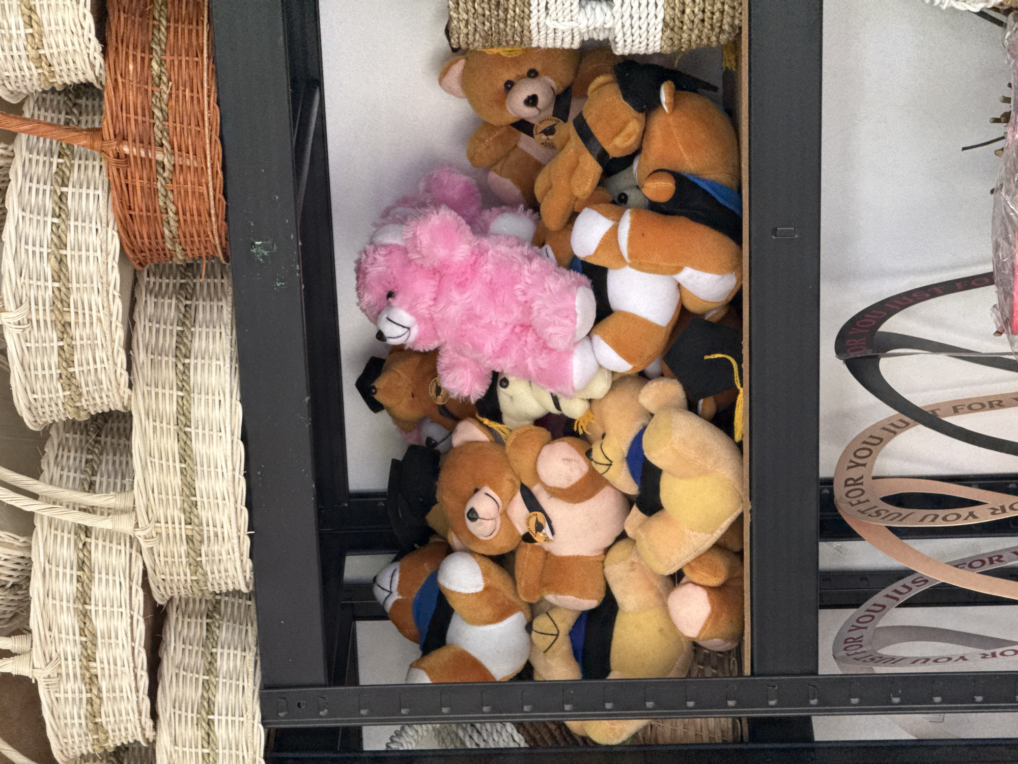 The image shows several shelves in a storage area. The top shelf displays an assortment of woven baskets in various sizes and colors, including natural straw, white, and a prominent orange basket. The middle shelf is filled with a collection of plush teddy bears. Some of the teddy bears are wearing graduation caps and sashes. A bright pink teddy bear is positioned prominently in the center of this shelf. The bottom shelf features several curved signs or decorative elements with the text "FOR YOU JUST FOR YOU" repeated on them. This storage area is likely part of a retail space or workshop located in Kuta Selatan, Indonesia, given the context of the request. The overall impression is one of organized storage of craft items and plush toys.
