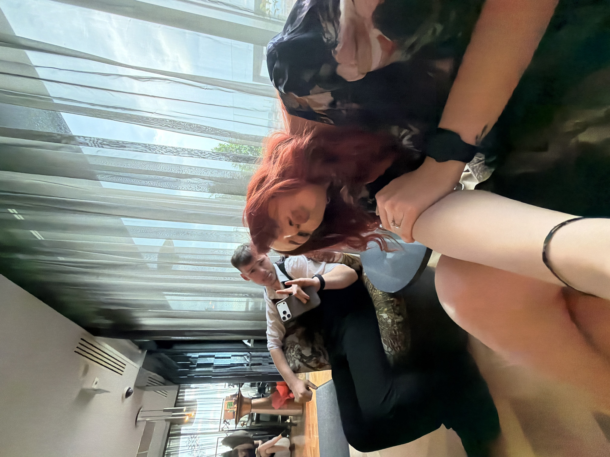 Two people are captured in an indoor setting, with a third person visible in the background. The location appears to be Sofia, Bulgaria. The red-haired person is holding the pale arm of the other, with visible black bands around the wrist. The background features a curtained window revealing a partial view of the outside, a ventilation system on the ceiling, and additional people in the distance. The second person is holding a phone, with their face is visible. The first and second people are positioned in a manner that suggests close interaction. The indoor setting appears to be a building with modern architecture.