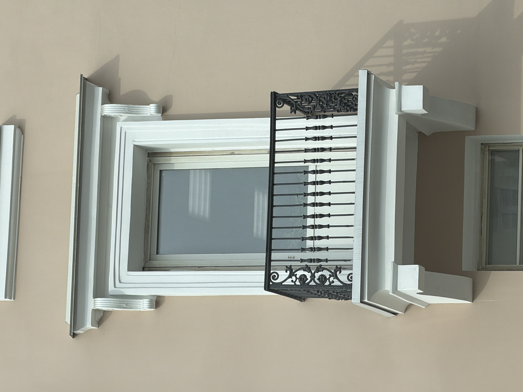 A window with ornate white molding and a decorative wrought iron grill, partially open, is set into a light beige wall. The window reflects light and shows a view of an interior space. The molding is multi-layered and includes decorative elements. The grill consists of vertical bars with decorative knobs and ornate scrollwork at the top and bottom. The wall appears to be exterior. The photograph was taken in Perm, Russia.