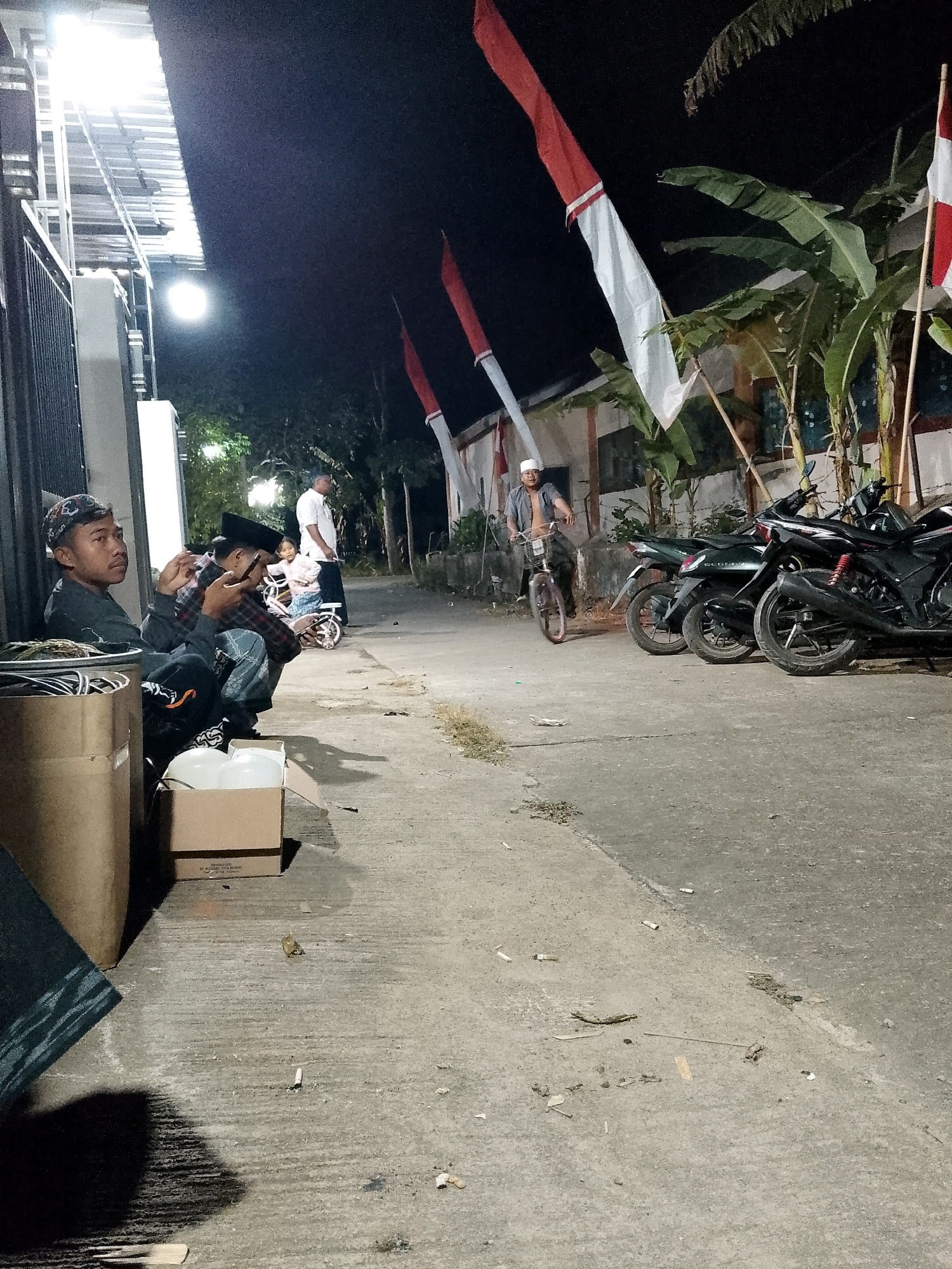 In Karangsari, Indonesia, at night, three men are seated on the ground near a corrugated metal awning and a fence. One man is looking to the right, another is looking at his phone, and the third is partially obscured. Behind them, a child is visible on a bicycle, and a man is standing. Further back, several motorcycles are parked. Indonesian flags are displayed on poles. The ground is paved concrete and shows signs of litter. A large cardboard box and a cylindrical container are also present.
