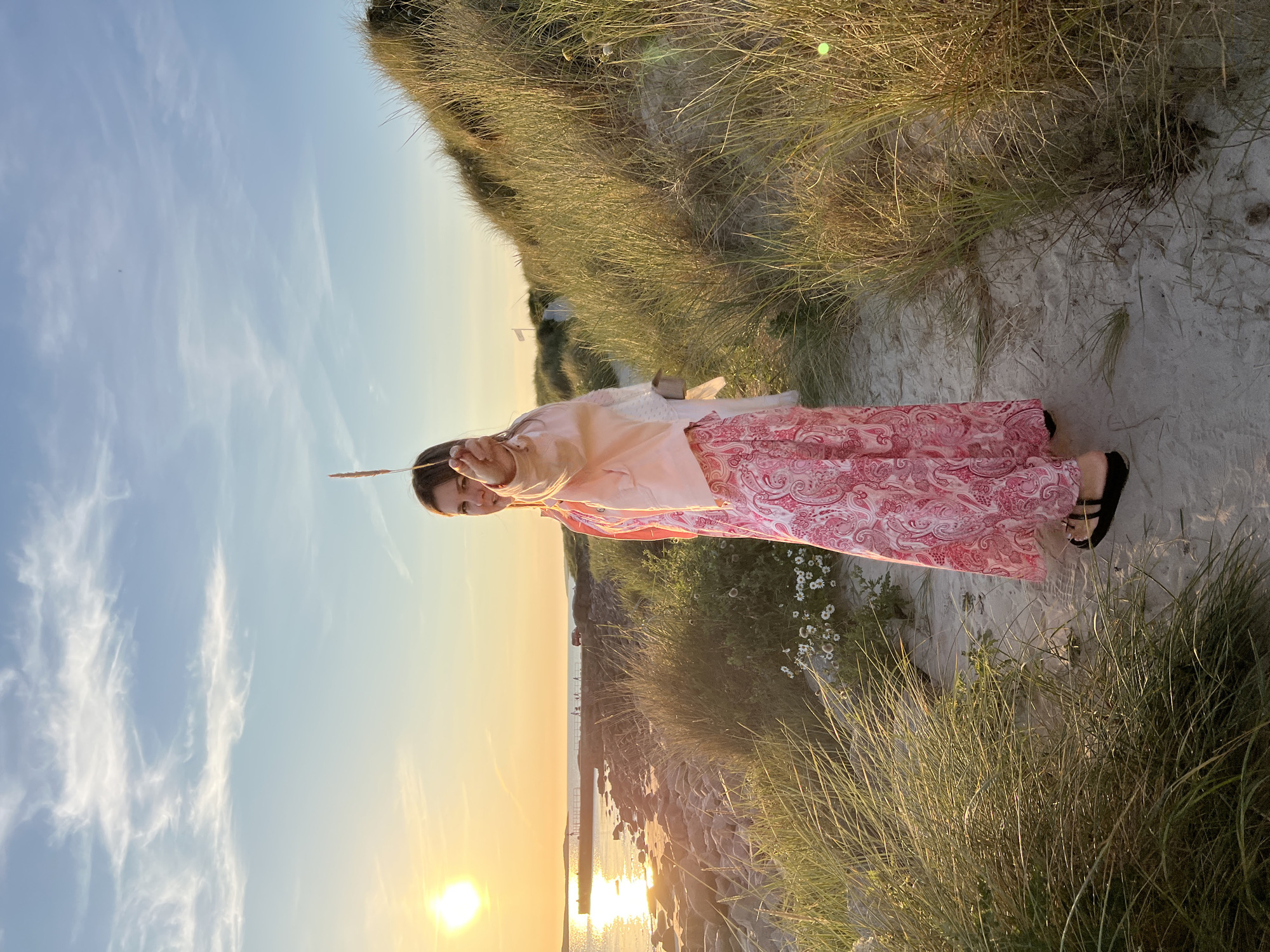 A woman stands on a sandy path through tall grasses on a coastline at sunset. She is wearing a long, pink paisley-patterned dress and a light pink jacket. She holds a stalk of grass in her outstretched hand. In the background, the sun sets over the water, casting a golden glow on the scene. A stone pier extends into the water, with several people and a vehicle visible on it. The sky is blue with wispy white clouds. This location is the coastline near Lunniaghbeg, Ireland.