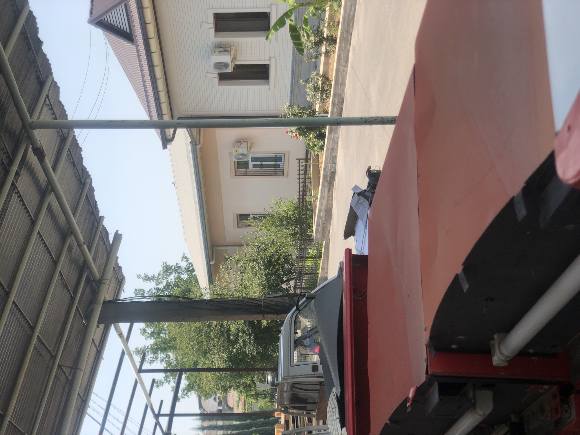 The image shows a street scene in Tashkent, Uzbekistan, with buildings on the right and a covered area with corrugated metal roofing in the foreground and left. A white pickup truck with a covered bed is parked under the structure. Several trees and bushes are visible, along with a metal fence. Two-story buildings with white and beige facades line the street, one with a dark brown roof and dormer window, and the other with a lighter colored roof. Air conditioning units are mounted on the exterior walls of the buildings. The ground appears to be paved. In the foreground, a large red industrial machine or vehicle is partially visible. The sky is clear and blue.