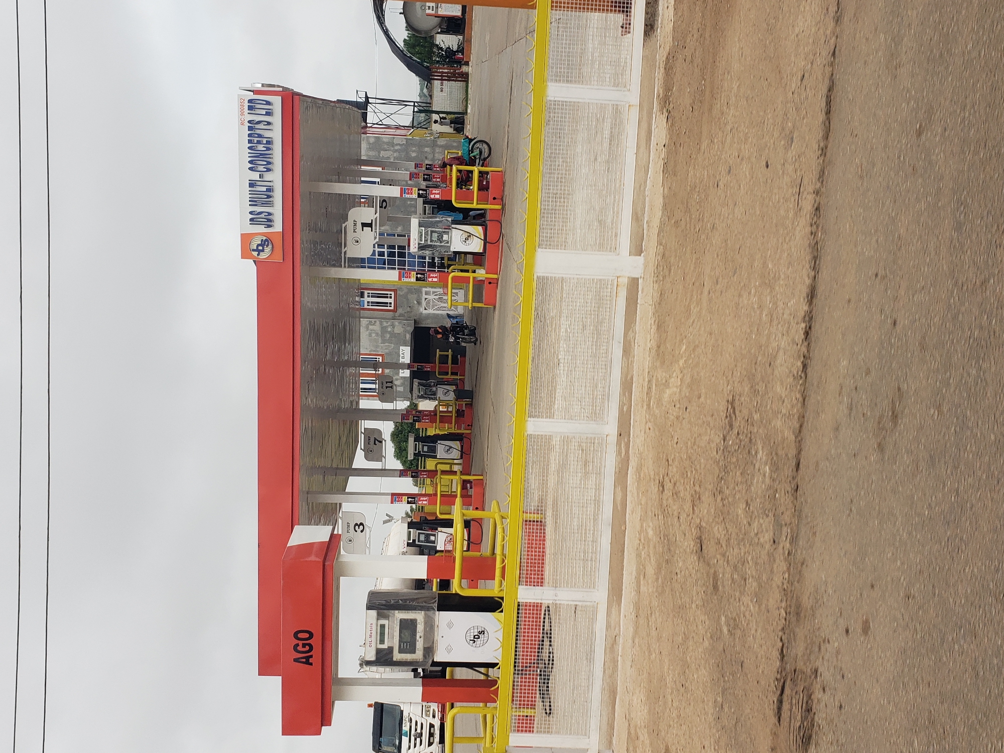 An outdoor, eye-level, medium shot captures a gas station under an overcast sky. The foreground features a dirt road and a white metal fence. Beyond the fence, a paved area of the gas station is visible, with several fuel pumps under a red and white canopy. The canopy is supported by white columns. The brand names "AGO" and "JDS MULTI-CONCEPTS LTD" are visible on the canopy. Numerous fuel pumps are arranged in rows, some labeled with numbers like "1" and "3". A white truck with "MNPC" visible on its side is parked to the left. Several people are present, some on motorcycles and some near the pumps. A person in a red shirt is standing at pump 1, and another person is on a motorcycle to the right. The environment appears to be dry and dusty.