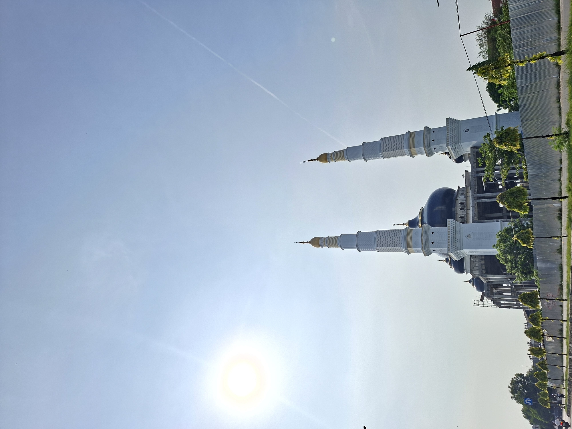 The image shows the Baiturrahman Grand Mosque in Pantegajah, Indonesia. The mosque features two tall, white minarets with golden tops, and a central dark blue dome. The foreground is dominated by a clear sky, with the sun visible near the bottom left. A vapor trail is also visible in the upper left corner of the sky. The mosque is bordered by a road with small trees planted along the median.