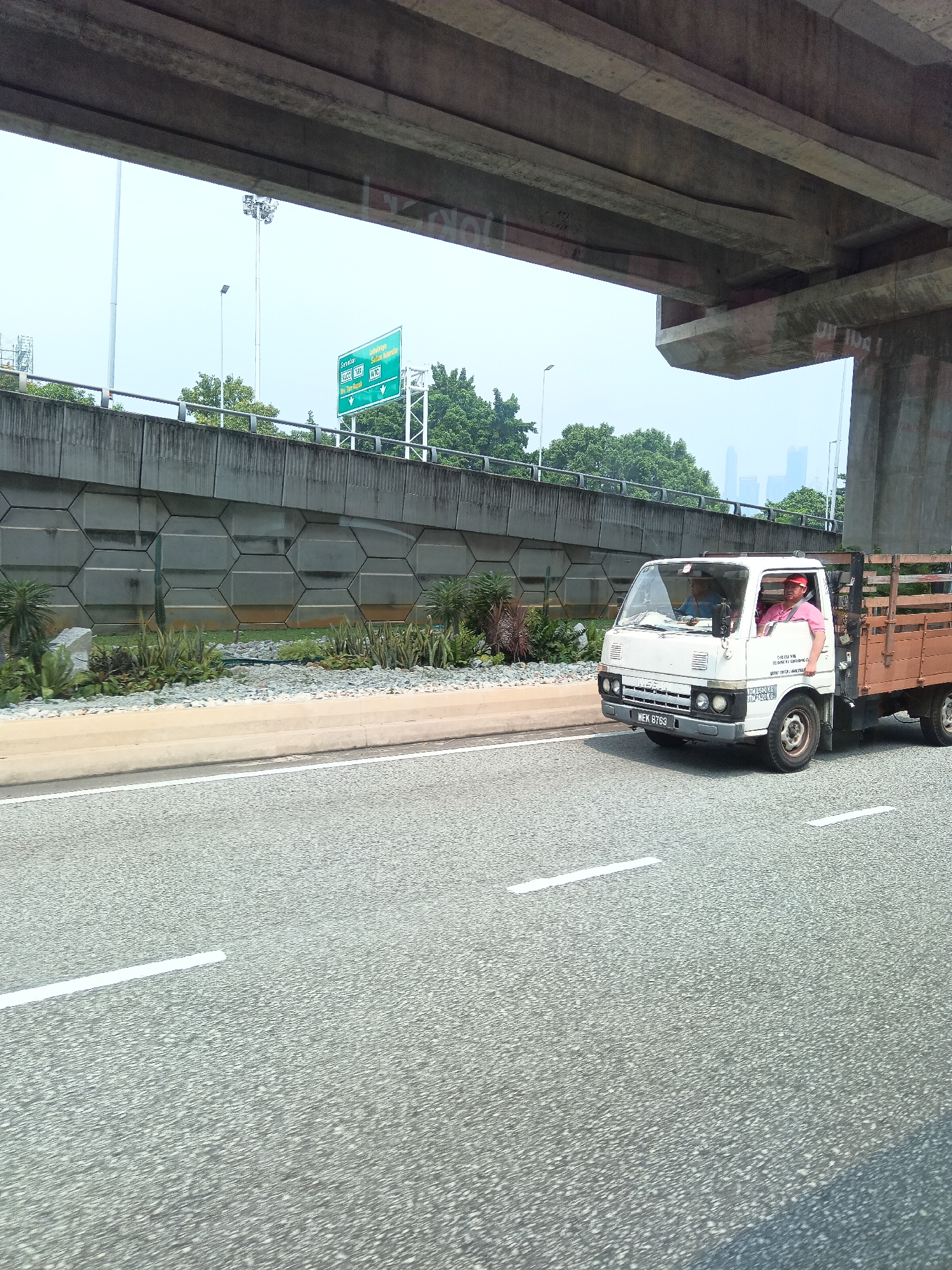 A white, flatbed truck with a wooden cargo area drives on an asphalt road under a concrete overpass in Kuala Lumpur, Malaysia. The truck has a license plate that reads "WEK 8763". Inside the cab, two men are visible. The driver, wearing a pink shirt and a red cap, is looking forward. The passenger, wearing a blue shirt, is looking down. On the side of the truck, text is printed. To the left of the road, a retaining wall made of hexagonal concrete blocks is visible, with some landscaping of rocks and plants. In the background, trees and distant buildings can be seen under a hazy sky. A green road sign pointing left and right is visible above the retaining wall.