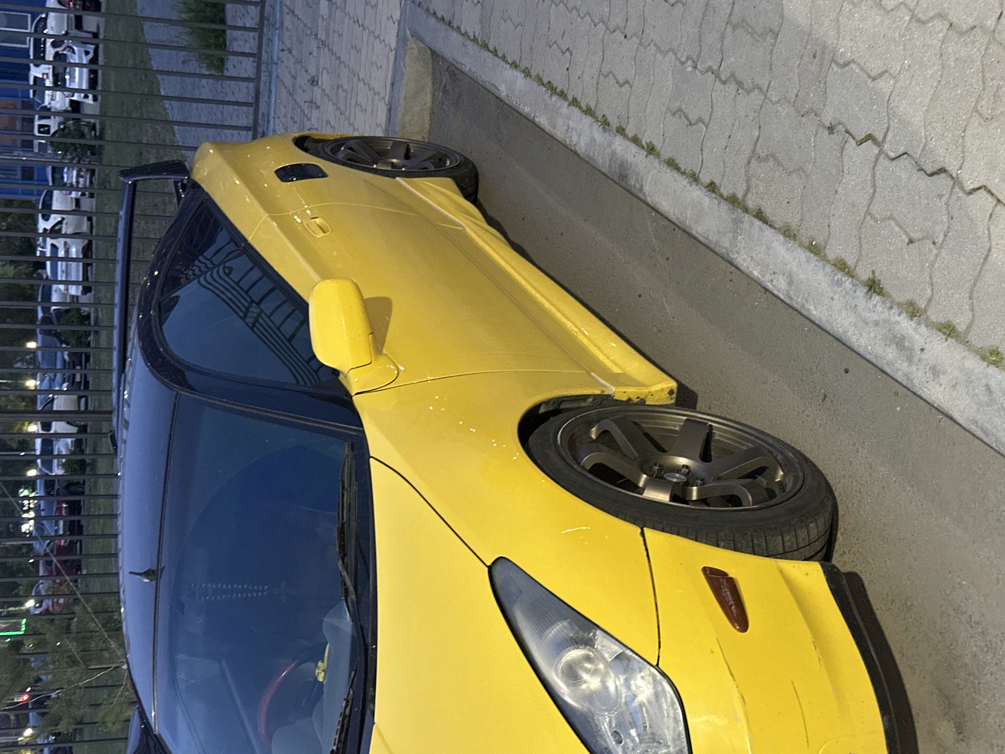 A bright yellow sports car, strongly resembling a 7th generation Toyota Celica (T230), is the central subject of the image, which is rotated 90 degrees clockwise. The car is parked outdoors, alongside a concrete curb and a sidewalk composed of grey interlocking concrete pavers with some weeds growing in the cracks.
The yellow car features custom dark grey multi-spoke wheels, with the front right wheel clearly visible. Its design includes clear headlight assemblies and an amber side marker on the front fender. A subtle black rear spoiler (ducktail style) is visible at the back, and the car's roof appears to be black.
In the background, behind a dark metal fence, several other vehicles, predominantly white, are partially visible, suggesting a parking area or dealership. Reflections of these vehicles are visible in the fence and the car's dark-tinted windows.
The scene is captured during dusk or nighttime, indicated by the low ambient light and the overall dark atmosphere. No specific weather conditions are discernable, and no human activity is depicted. There is no visible text within the image. The provided context indicates the location is Khabarovsk, Russia.