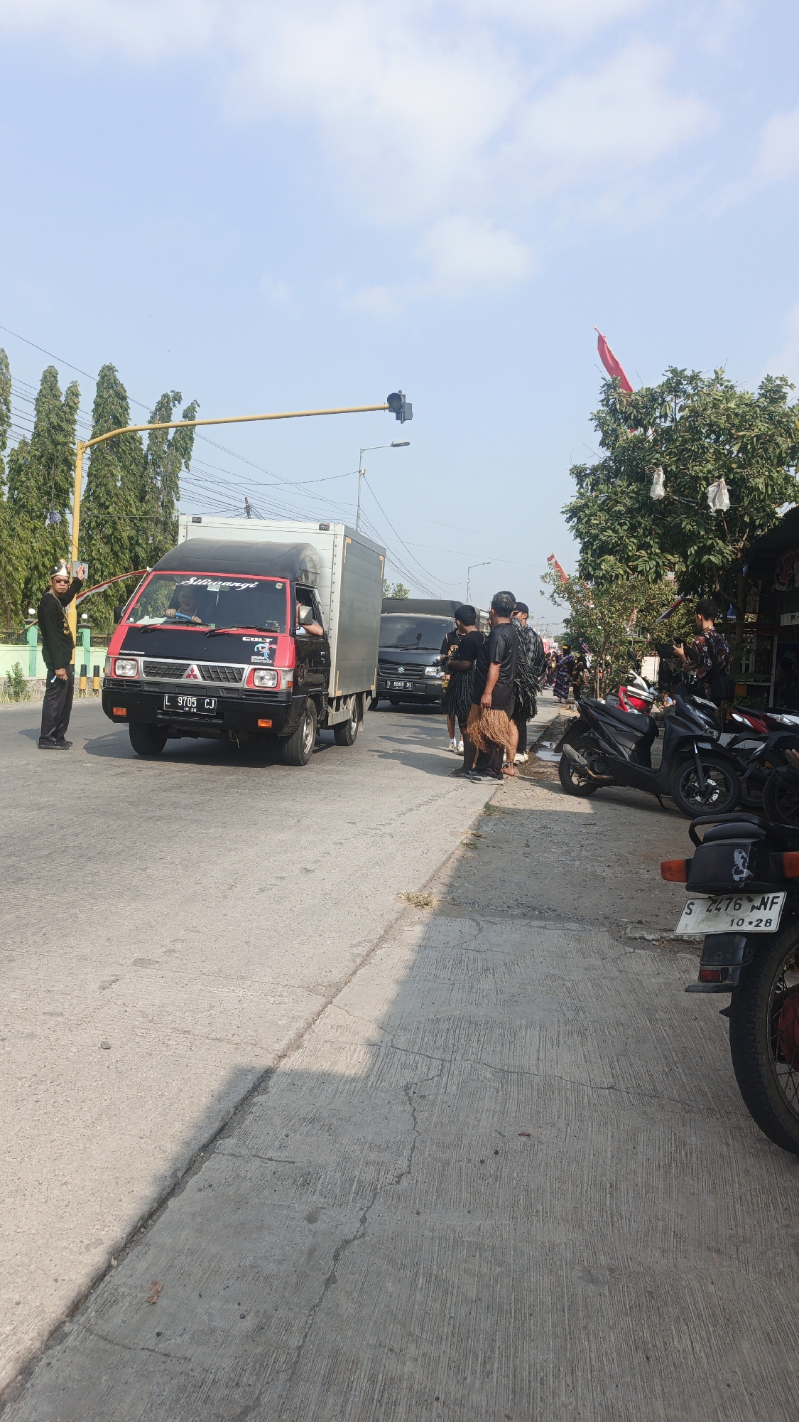 The image depicts a street scene in Sumengko, Indonesia, under a clear sky. A red and black truck with a silver cargo box is parked on the right side of the road, facing left. A man in a black uniform with a white hat and a white sash is standing on the left side of the road, facing the truck and holding his hand up. Several people in black clothing are gathered on the sidewalk near the truck, some holding what appears to be a traditional woven skirt or mat. A black van is parked behind the truck. To the right of the road, several motorcycles are parked along a sidewalk that leads to shops. A red flag is visible on a tree in the background. The road surface is concrete. The overall impression is of a gathering or event taking place on a street.