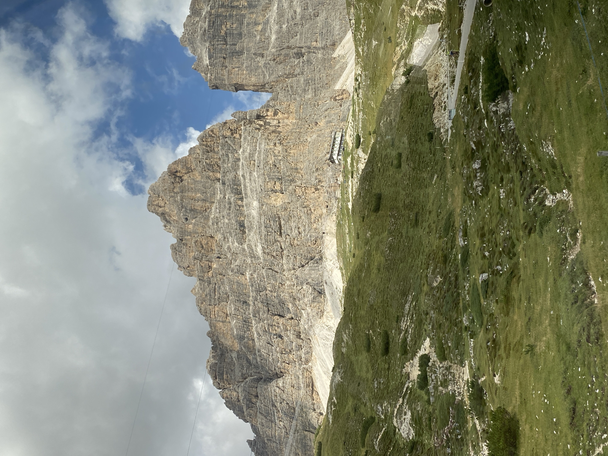 This is a landscape photograph of the Tre Cime di Lavaredo, a striking mountain formation in the Dolomites mountain range, located in the Veneto region of Italy, near the municipality of Auronzo di Cadore. The image captures a vast, rugged mountain face with distinct geological layers of grey and brown rock. A building, appearing to be a rifugio or mountain hut, is situated on the lower slopes of the mountain, nestled amongst the green, grassy terrain. The foreground consists of a verdant, undulating hillside dotted with rocks and sparse vegetation, with a winding road visible in the lower right corner. The sky above is partly cloudy, with patches of blue sky peeking through. In the upper left portion of the image, a pair of thin, dark lines are visible, suggesting a cable or wire, possibly from a ski lift or cable car system.