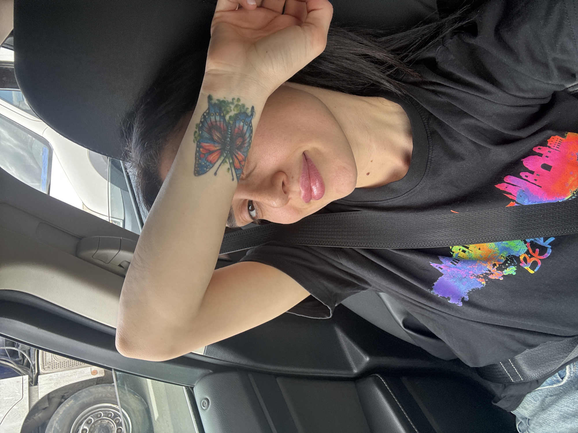 A woman is pictured inside a vehicle, likely a car or truck, with a seatbelt visible across her chest. She is wearing a black t-shirt with a colorful design on the front. She has dark hair and is wearing a wrist tattoo of a butterfly. The location of the city of Tbilisi, Georgia cannot be determined from this image.
