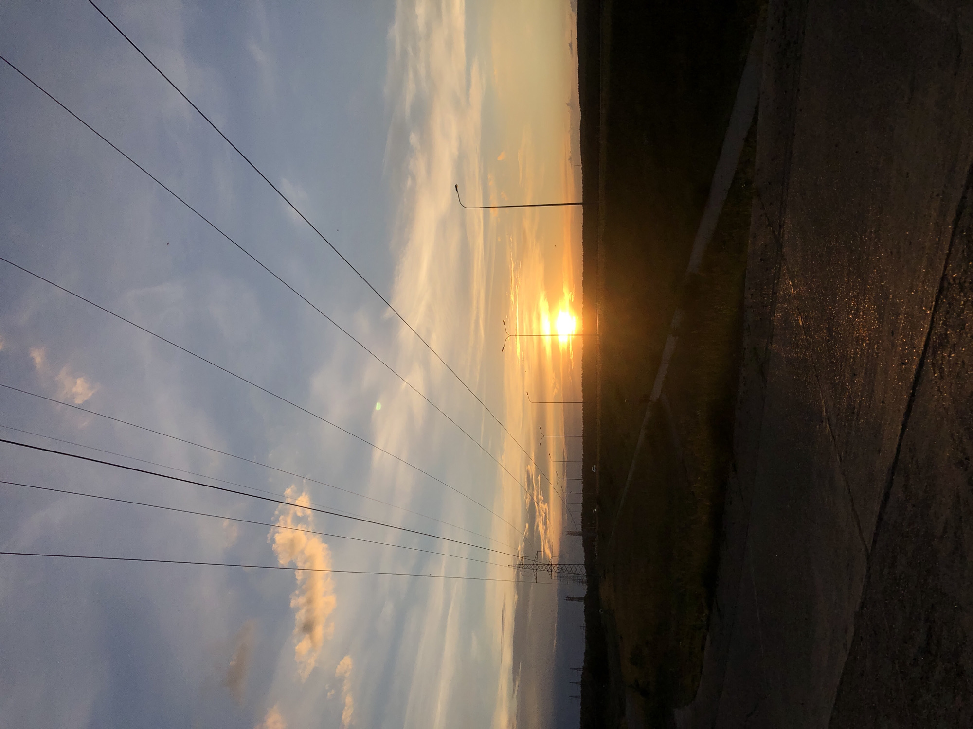 A twilight photograph captures a rural landscape with a paved road in the foreground and a sunset in the background. Power lines crisscross the sky, with tall electrical towers visible in the distance. The sun, a bright orb, is setting on the horizon, casting a warm glow across the sky and illuminating some clouds. Streetlights line a road that curves into the distance. The scene is devoid of any people or animals. The image appears to be taken in a location outside of the city of Chaikovsky, Russia, given the rural characteristics of the landscape and absence of urban infrastructure typically found within a city.