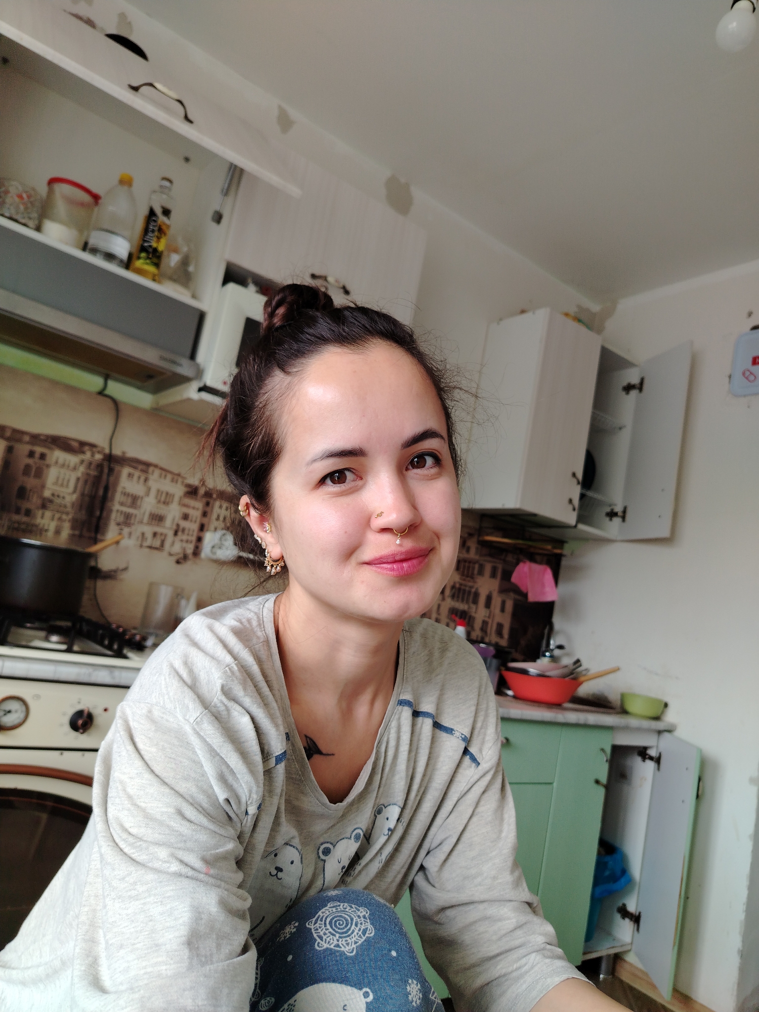 A female individual is depicted in a domestic kitchen located in Noviy, Russia. The woman, with dark hair tied in a messy bun, is smiling and looking directly at the camera. She has a medium complexion, brown eyes, and wears light pink lipstick. Her left nostril features a small gold stud, and she has a gold septum ring with a dangling pearl-like bead. Multiple gold earrings, including hoops, studs, and decorative dangling elements, are visible on both ears. She is wearing a light grey long-sleeved t-shirt with a polar bear graphic and blue accents on the shoulders, paired with blue pants featuring a white circular pattern. A small dark bird tattoo is visible on her upper left chest.
The kitchen environment includes white walls with areas of minor damage or peeling. White upper cabinets are installed, with some doors open, revealing bottles (including "Olien" brand oil) and containers. A silver range hood is positioned above an off-white gas stove with visible control knobs, upon which a black pot with a wooden spoon rests. The backsplash features a sepia-toned cityscape mural. Mint green lower cabinets are visible on the right, with one door open to reveal a blue bag inside. A light-colored countertop holds a red mixing bowl, a green bowl, and various kitchen utensils. An exposed light bulb hangs from the white ceiling in the upper right corner of the frame.