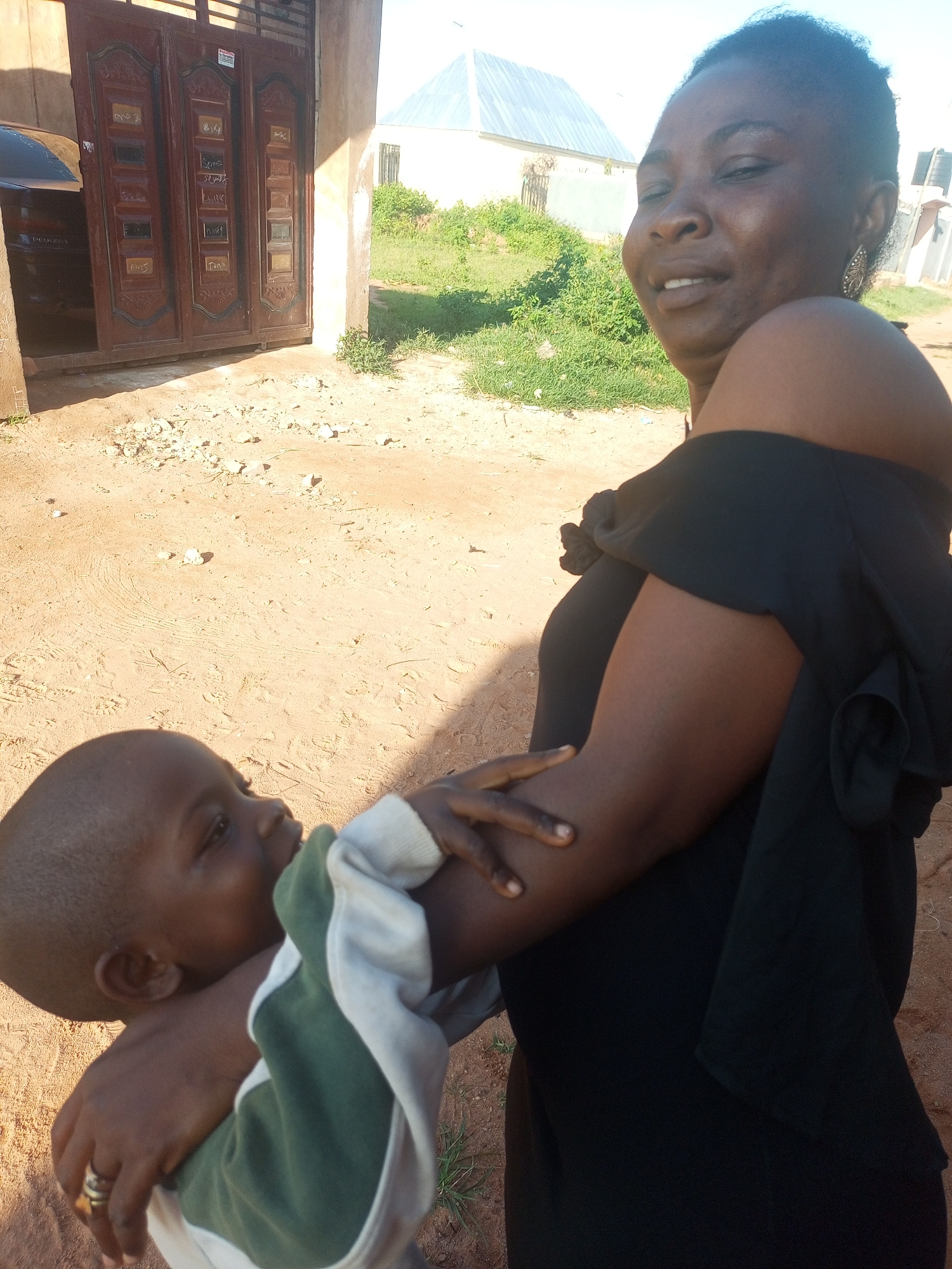 A medium shot captures a woman and a young child in an outdoor environment in Tempure, Nigeria. The woman, with her head tilted back and eyes closed, is holding the child in her arms. She is wearing a black off-the-shoulder top and a prominent silver earring in her left ear. The child, facing left and looking downwards, is dressed in a green and white garment. The background reveals a sandy ground with scattered small stones and sparse grass. To the left, a large, brown, ornate double gate with vertical panels is visible, partially obscuring a dark vehicle. Behind the gate and to the right, a light-colored building with a corrugated metal roof is situated, surrounded by green vegetation and more light-colored structures. The scene is illuminated by natural daylight, casting shadows on the ground.