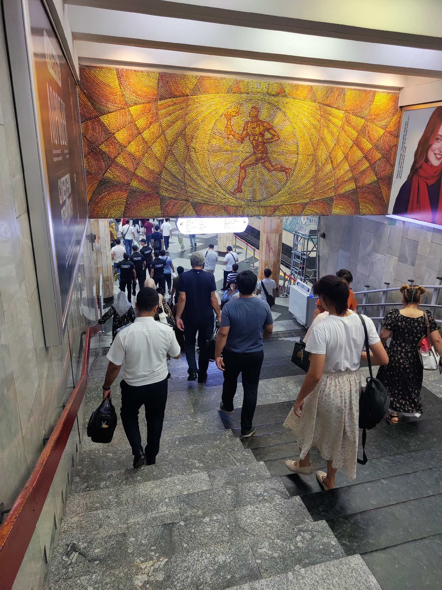This is a medium-angle shot taken from an elevated position, looking down a wide, well-lit staircase. The staircase is constructed with grey granite or similar stone, featuring a red-painted metal handrail on the left side. At the top of the stairs, a large, colorful mosaic artwork adorns the wall, depicting a stylized runner holding a torch within a sun-like radial pattern. To the right of the mosaic, a large advertisement featuring a smiling woman with red hair and a red scarf is visible.
A crowd of people are descending the stairs. In the foreground, a man in a white shirt and black trousers carries a black bag. To his right, a woman in a white dress with a floral pattern and espadrille shoes also descends. Behind them, several individuals are walking down, including a woman in a black floral dress. Further up the stairs, a larger group of people, predominantly men in black t-shirts with white lettering and others in casual attire, are also moving downwards. The environment appears to be an underground passage or metro station entrance in Tashkent, Uzbekistan, given the mosaic style and advertising common in such locations. The lighting is bright and appears to be artificial.