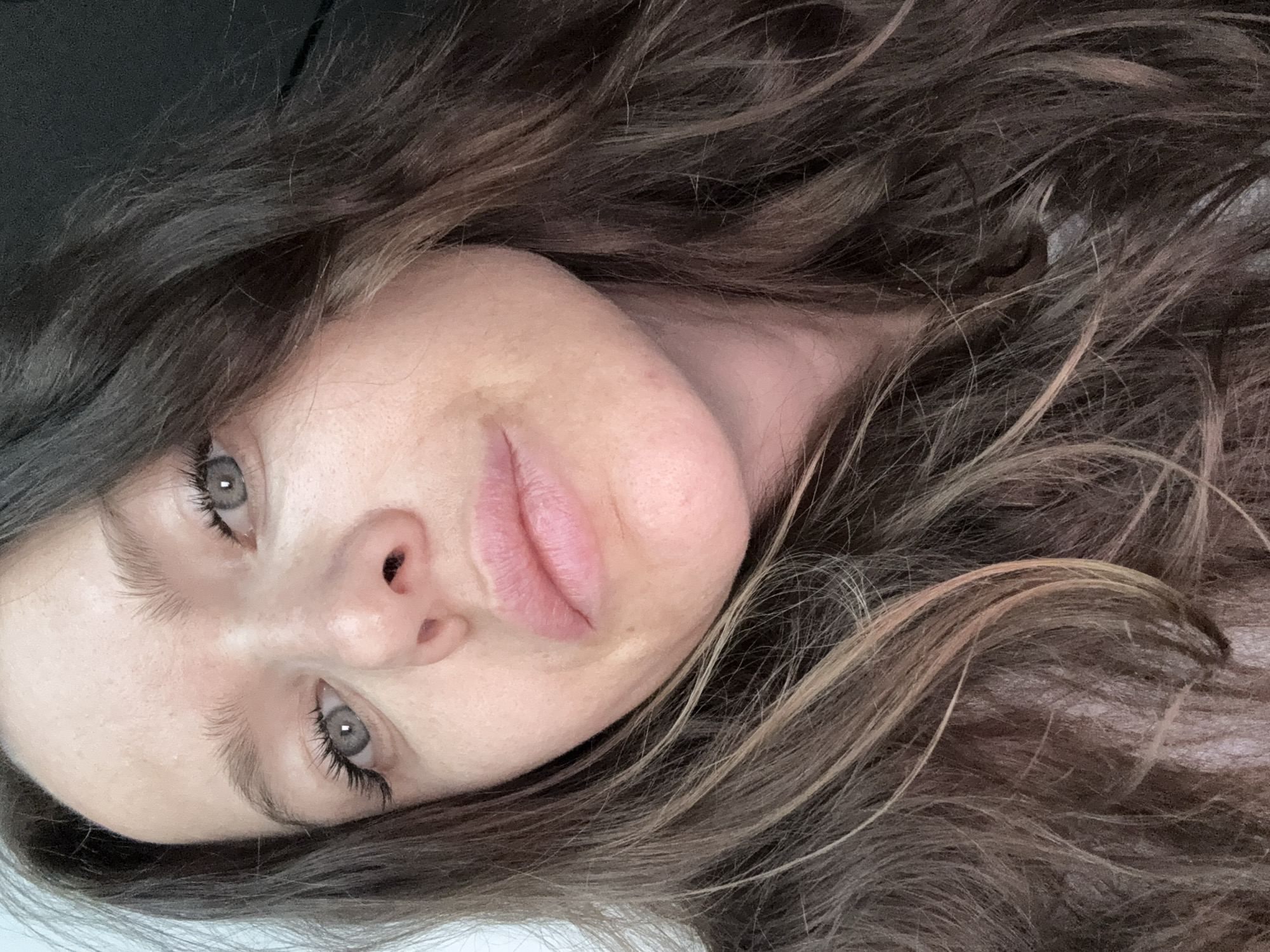 A close-up shot of a woman's face, tilted to the left. She has light skin, grey eyes, and brown hair with blonde highlights. Her lips are slightly parted. The background is blurred and indistinct, appearing dark on the top left and lighter on the bottom right. The location is unknown, and there is no indication of it being in Dolgoprudny, Russia.