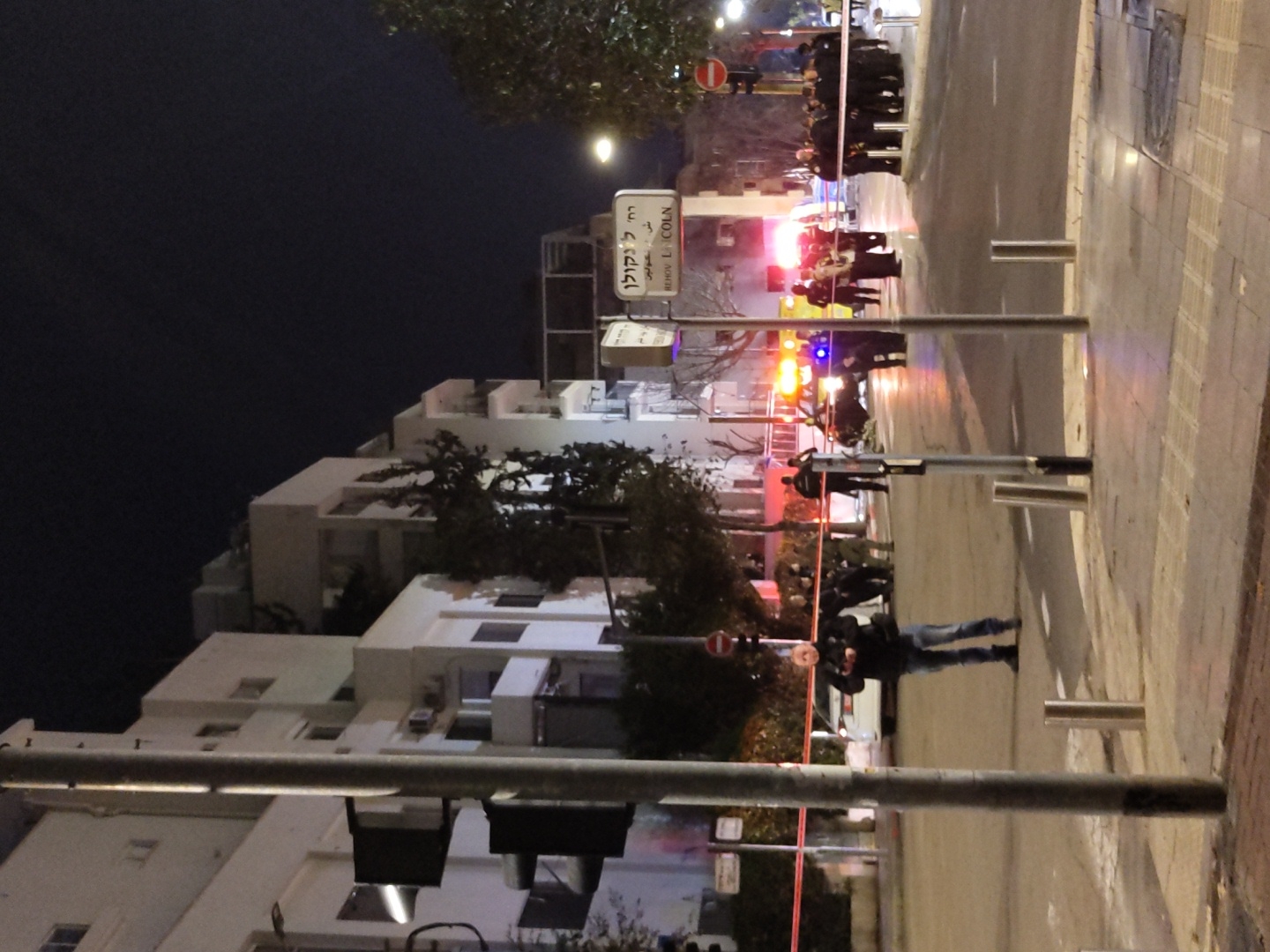 An urban street intersection at night, identified by a street sign as Rehov Lincoln (רחוב לינקולן) in Tel-Aviv, Israel, shows evidence of an ongoing incident. A red barrier tape spans the roadway, sectioning off an area. On the far side of the tape, multiple emergency vehicles are present, indicated by prominent flashing blue and red lights, illuminating a cluster of individuals. A dark-colored vehicle is visible near the flashing lights, and a white sedan is parked behind the tape. A large group of approximately 10-15 individuals, many in dark uniforms consistent with police or emergency services, are gathered on the right side of the street near the flashing lights, some appearing to be in discussion. Another group of uniformed personnel is visible further down the street. In the immediate foreground, a civilian man in a dark jacket and blue jeans stands on the sidewalk, holding a small dark object, possibly a phone or camera. Traffic light poles and several metal bollards line the sidewalk. Multi-story white residential buildings with balconies and trees are visible in the background under a dark night sky.