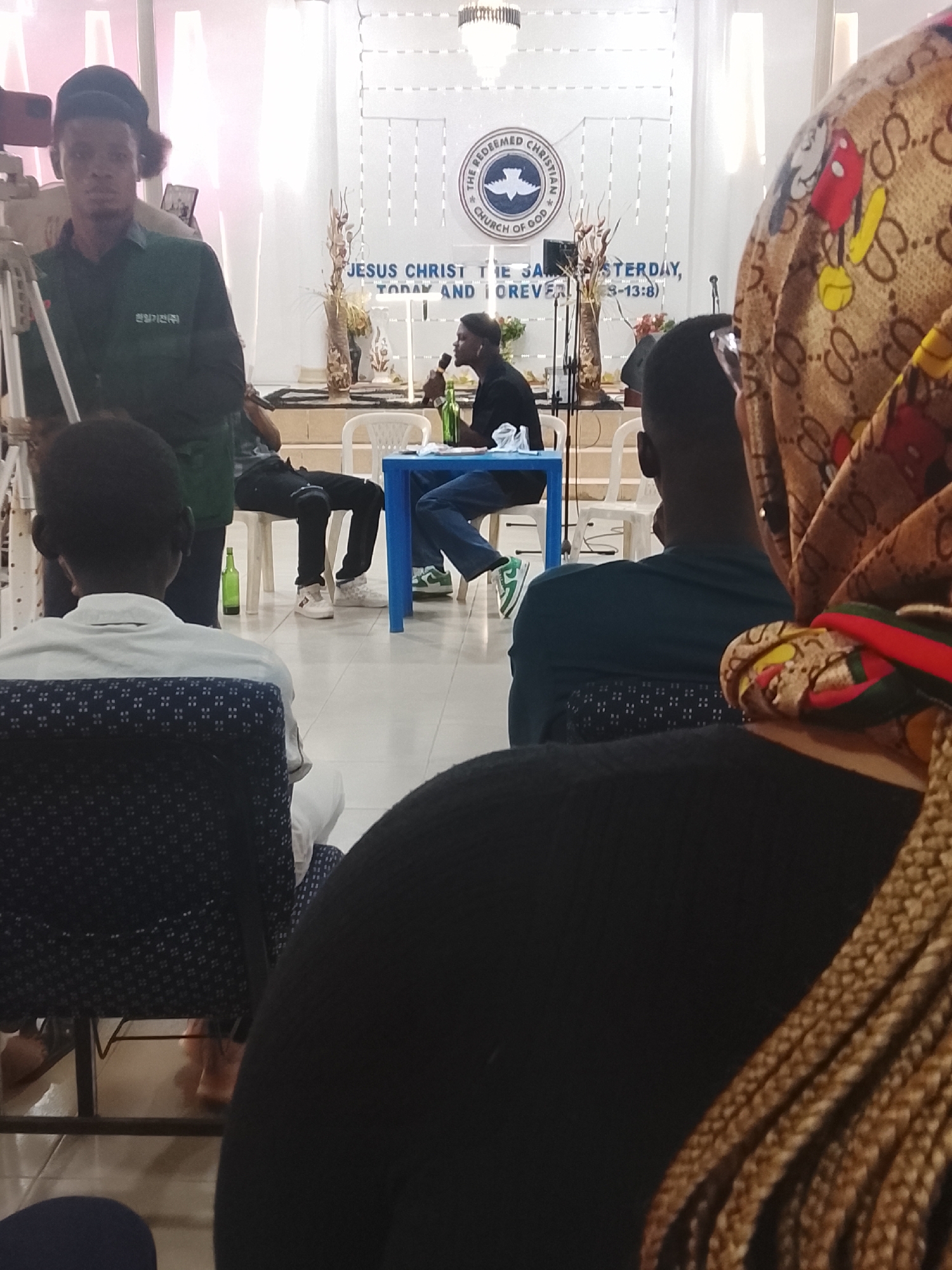 The image shows a gathering indoors, possibly in Jos, Nigeria, based on the church inscription visible in the background. A man is seated at a blue table speaking into a microphone. A green bottle is on the table. Another man sits to the side, casually posed in a chair. A cameraman stands behind a tripod. There are people seated in the foreground, with their backs to the camera. A woman with patterned headwear and braided hair is prominent on the right. In the background, the logo of "THE REDEEMED CHRISTIAN CHURCH OF GOD" is visible above the words "JESUS CHRIST THE SAME YESTERDAY, TODAY AND FOREVER (3-13:8)". The room is brightly lit.