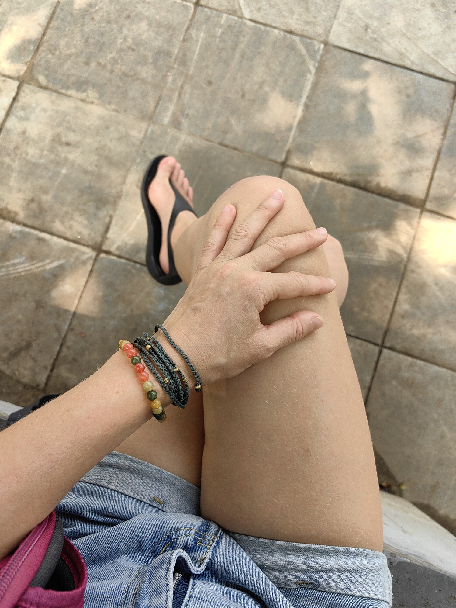 A person is seated outdoors on a tiled surface. The individual is wearing denim shorts, a pink bag, and black flip-flops. They have bracelets on their left wrist. Their left hand rests on their right knee. The location is indicated as Thành phố Hà Giang, Vietnam.