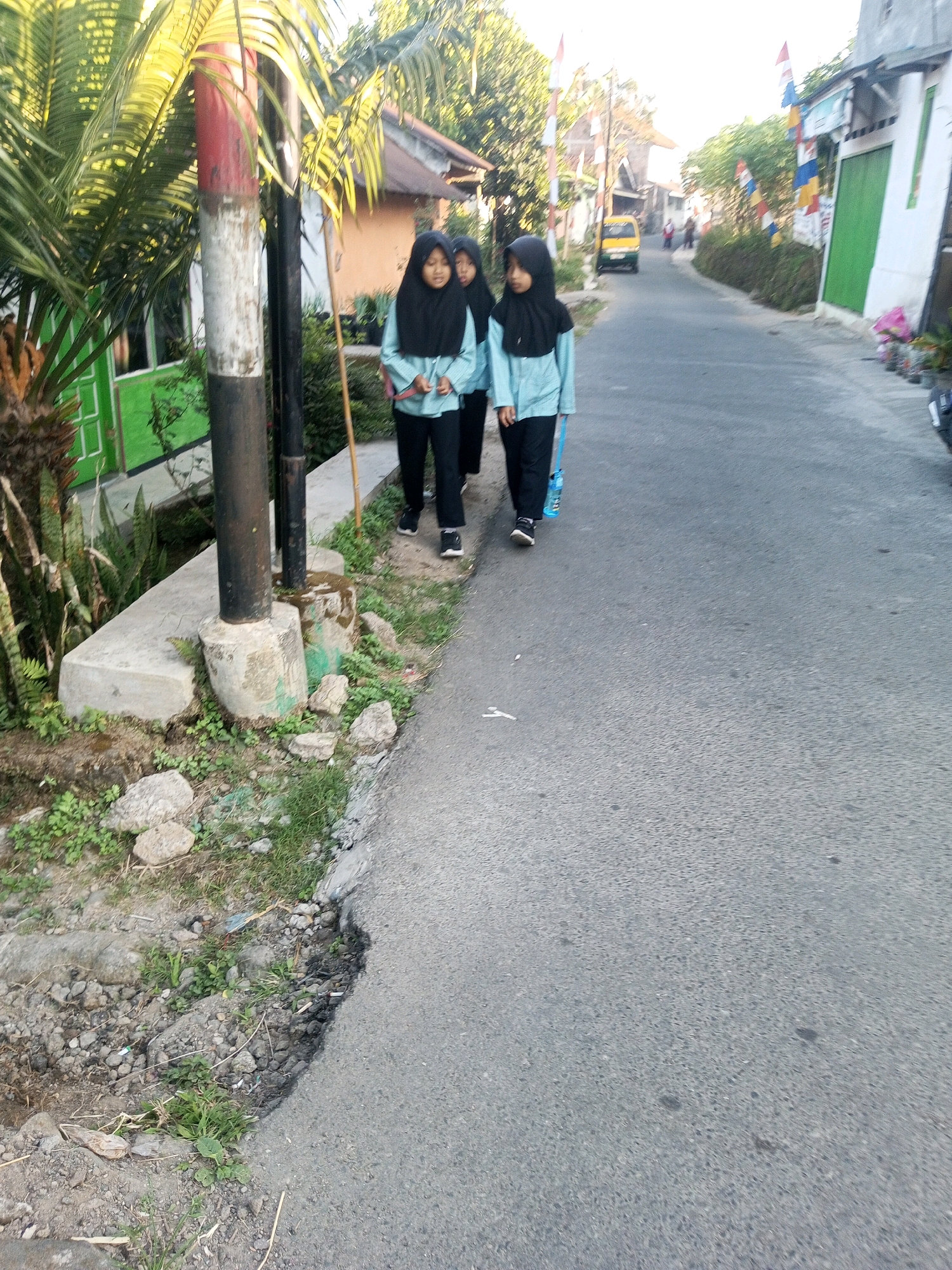 Three young girls, dressed in light blue tops and black headscarves, walk down a paved road in Andongsili, Indonesia. The girl in the foreground is looking down and holding a blue water bottle. The girl behind her is looking forward, and the third girl is mostly obscured by the other two. Sunlight casts long shadows across the road, indicating it is late afternoon or early morning. The road is flanked by houses and vegetation, with a yellow van visible in the distance. A red and white striped pole stands to the left of the frame, near a palm tree. The road's edge is uneven, with dirt and rocks visible.