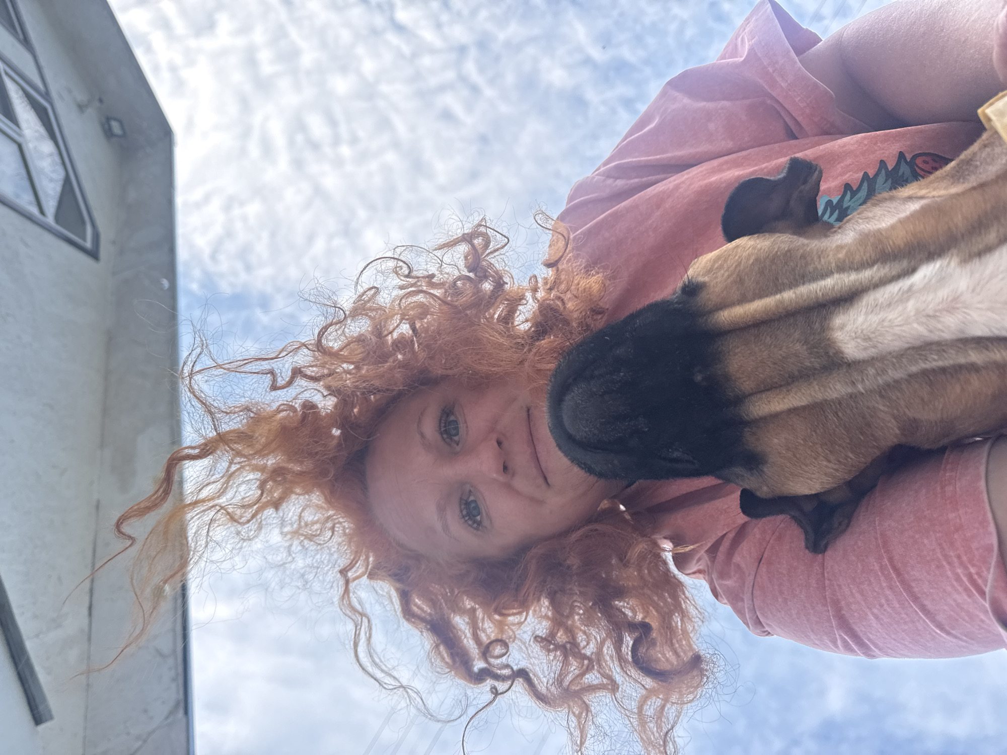 A person with red curly hair, fair skin, and blue eyes is holding a brown and black dog. The dog's nose is pressed against the person's face. The person is wearing a pink t-shirt. The sky is visible in the background, with white clouds. The corner of a white building with a window is visible on the left side. There is not enough information in this image to determine the exact location in Chaikovsky, Russia.