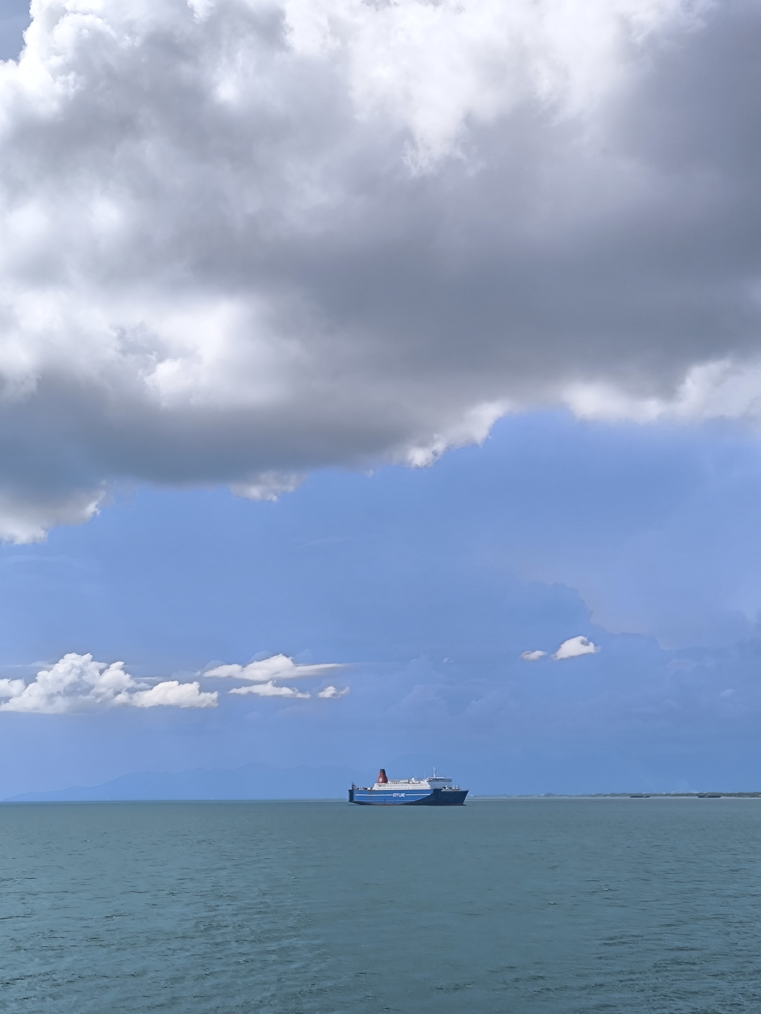 A large passenger and vehicle ferry, featuring a blue hull, white superstructure, and a red stripe near its funnel, cruises across a vast body of calm, teal-colored water. The ship is positioned slightly off-center to the right, moving from left to right across the frame.
The sky above is dramatic and multi-layered, dominated by a mix of heavy, dark grey clouds and brighter, fluffy white cumulus clouds. Patches of clear, vibrant blue sky are visible between the cloud masses, particularly in the mid-ground. The contrast between the dark and light clouds suggests dynamic weather conditions, possibly leading to or following rain.
Distant land masses are faintly visible along the horizon, especially on the left side, appearing as subtle blue-grey silhouettes against the sky and water. This implies an open sea or strait with nearby islands or coastlines, consistent with the location of Lembar Selatan, Indonesia.
The overall scene conveys a sense of peaceful transit amidst grand, changeable weather. The time of day appears to be midday, with diffuse sunlight filtering through the cloud cover. On the side of the ferry's blue hull, the white text "CITYLINE" is clearly discernible.