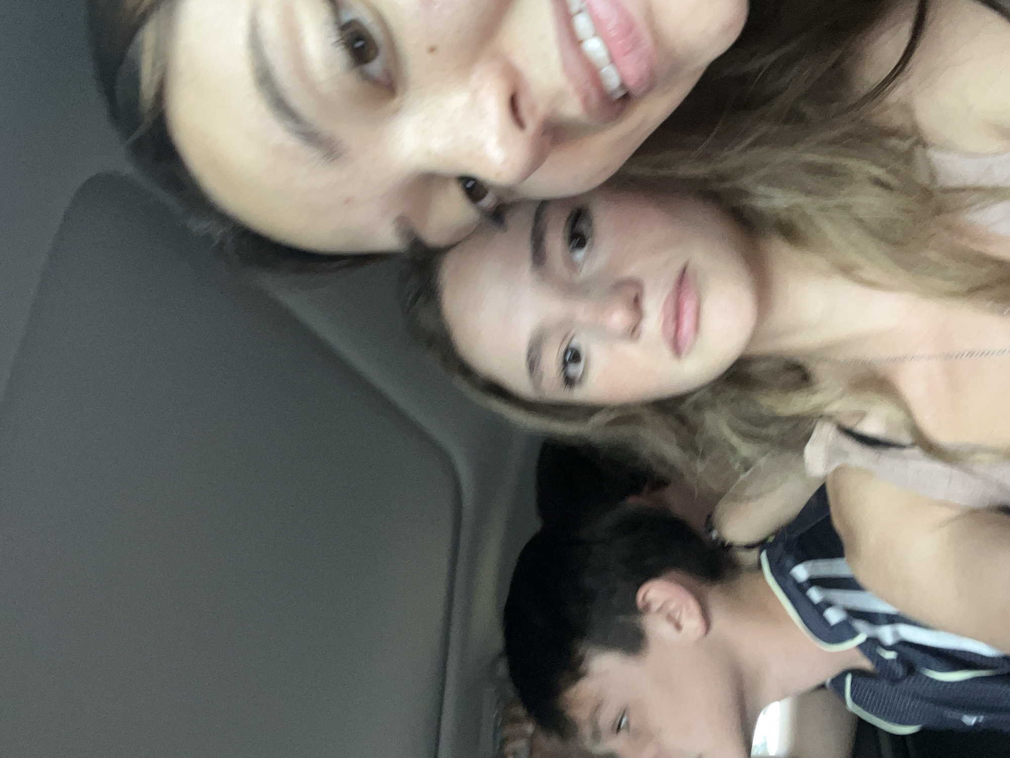 Three individuals are in the scene. Two women appear to be closer to the camera and positioned above the third person. The top woman has dark hair and is angled downward toward the camera. The middle woman has blonde hair and is looking at the camera. The person in the background has dark hair and is wearing a dark shirt with white stripes. The setting seems to be within a vehicle, given the interior paneling in the background. There is no specific indication of San Bernardino Tlaxcalancingo, Mexico.