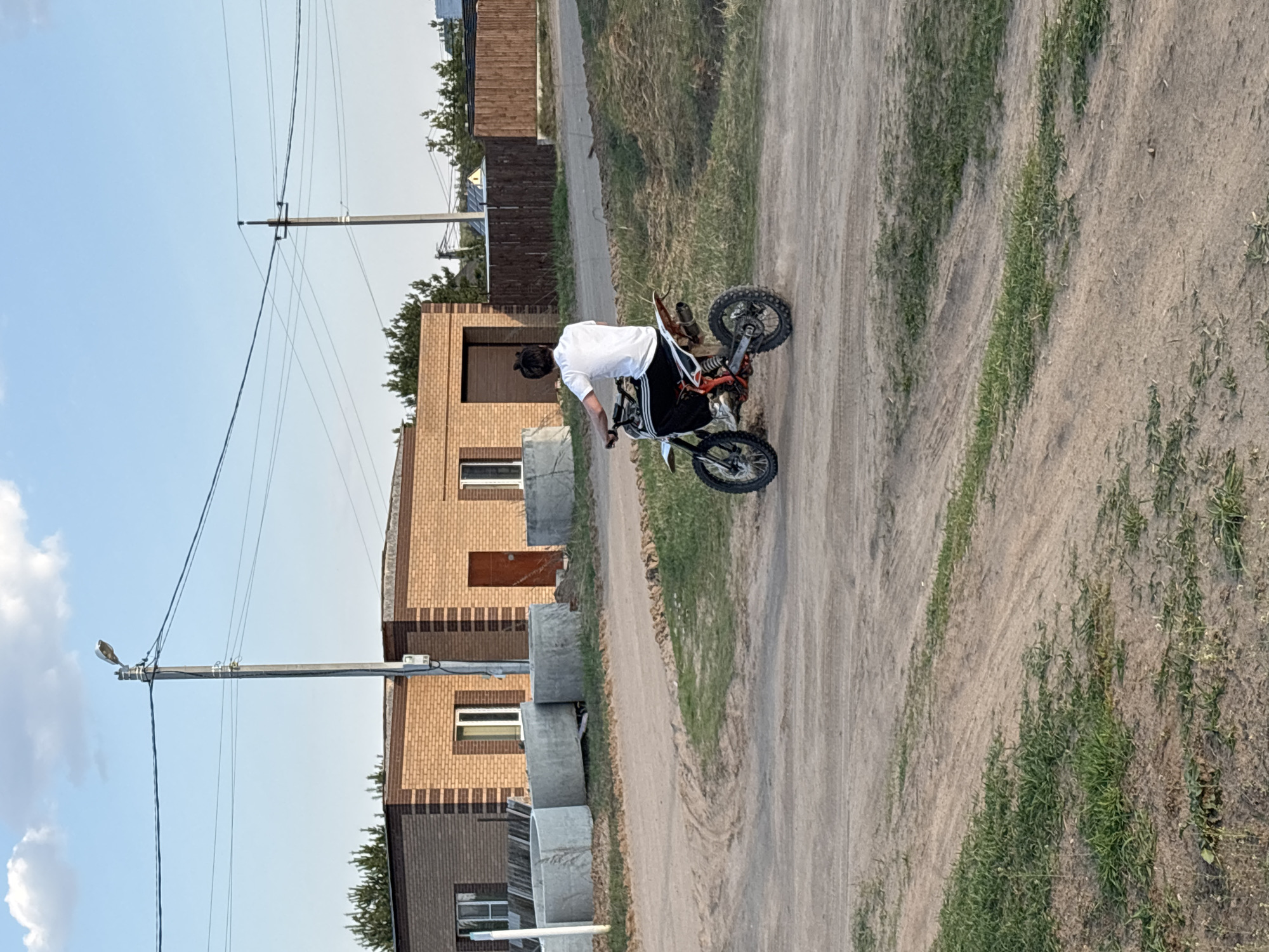 A person is on a dirt bike on a dirt road. The person is wearing a white shirt and dark pants. The bike is orange, black and white. The surrounding area is a rural neighborhood with houses and utility poles. The location is most likely the city of Chaikovsky, Russia.