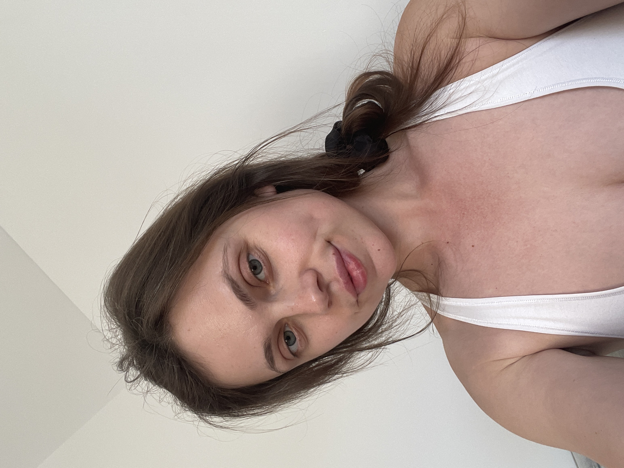 A young woman with brown hair and blue eyes is photographed from the chest up in what appears to be an indoor setting against a plain white wall. She is wearing a white tank top and her hair is pulled back with a black scrunchie, with some strands loose around her face. Her expression is neutral, with a slight smile. The lighting is natural and appears to be coming from the side, casting soft shadows. The image is a close-up, focusing on her face and upper body. There is no discernible context suggesting the location is in Szklarska Poręba, Poland.