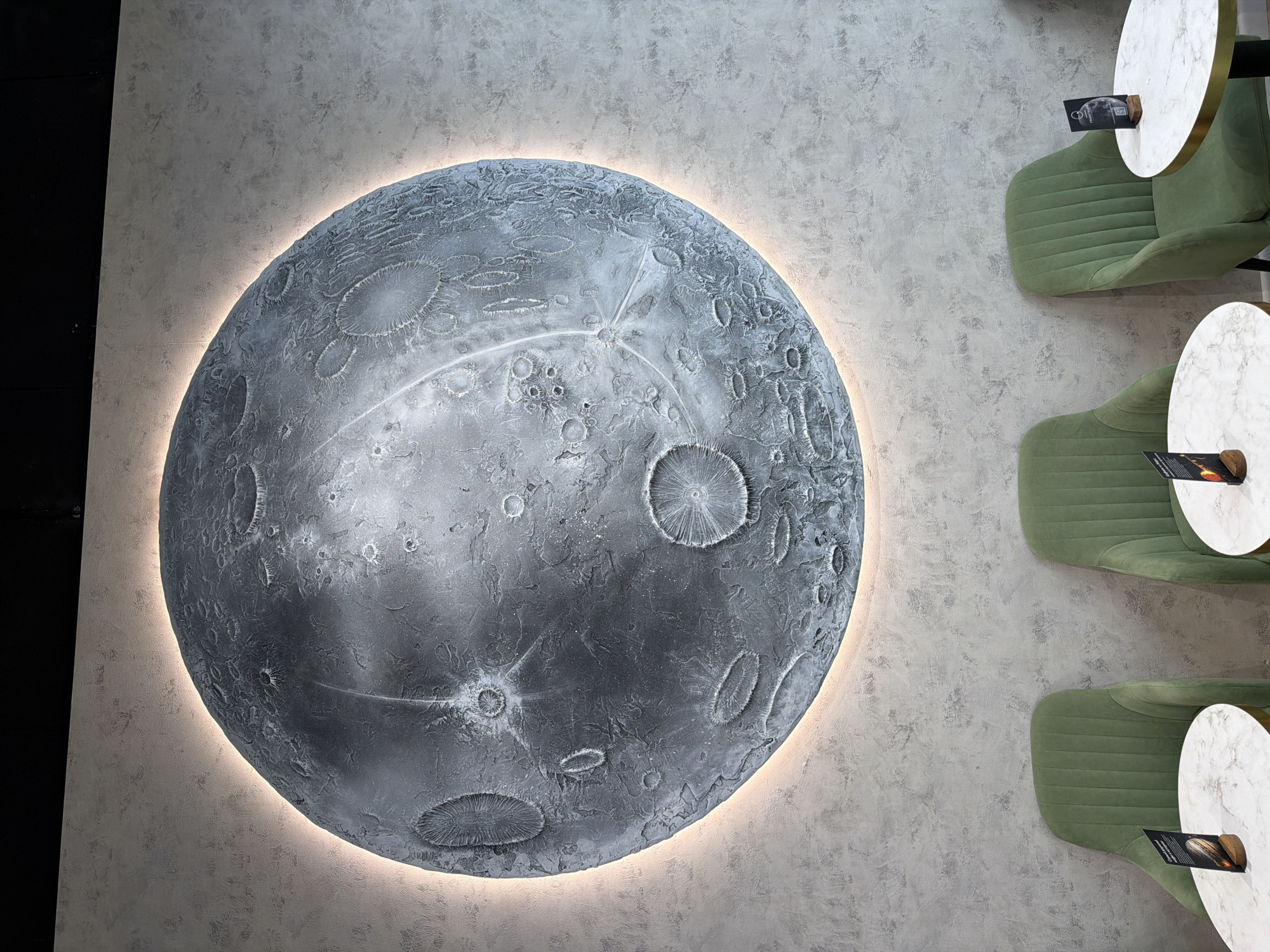 The image depicts an indoor setting with a large, circular, three-dimensional art piece resembling the moon affixed to a light gray textured wall. The moon's surface is detailed with craters and ridges, illuminated by a soft, ambient light from behind. To the right of the moon art, three green, upholstered chairs are arranged sequentially. Each chair has a small, round table with a marble top next to it. The bottom left corner of the image shows a dark, vertical surface, possibly a wall or panel. The location is Unknown, Unknown.