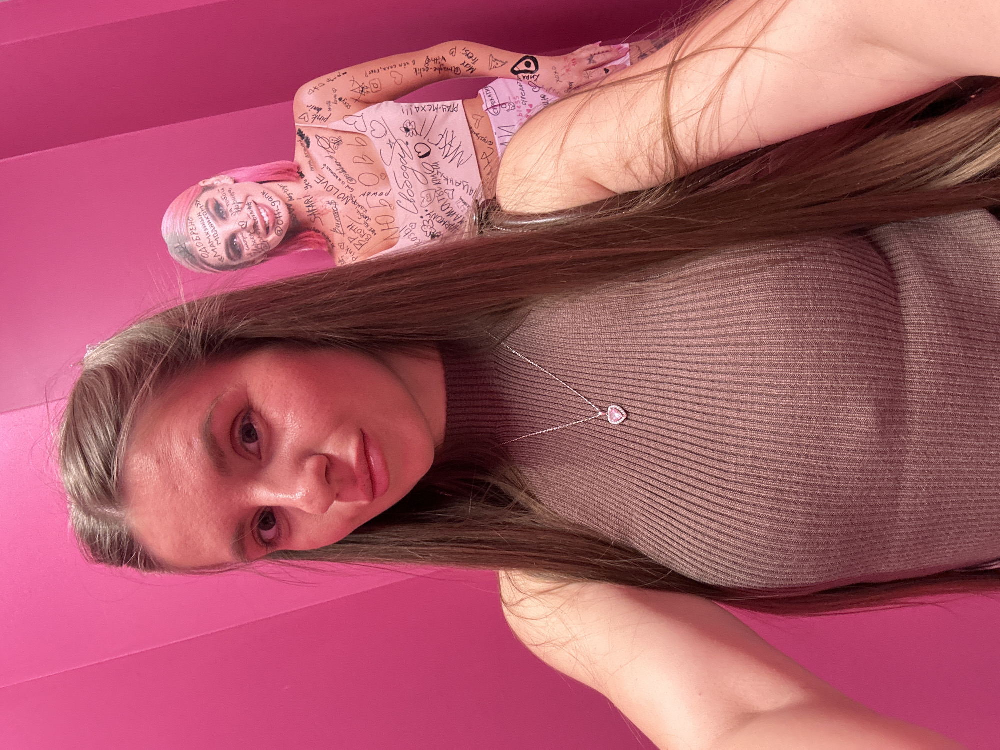 A selfie taken by a light-skinned woman with brown hair and wearing a brown ribbed sweater and a necklace with a heart-shaped pendant. Behind her is a pink wall with a cutout of a pink-haired woman in a pink outfit covered in writing. The writing on the wall includes the phrases "ОДОБРЕНО «МАМКИНЫН модником»", "Смотри за тобой @Offi-gank", "SAFARI You", "Pink BKTRO", "Ртуть лучший @rochkand", "Свобода", "HARMONY C", "чататы @ryzys", "sexy bair pink", "PΓAY-MCXA!!!", "May Amagbe-gelik", "Treysi B", "В чём СКЛА, БРАТ?", "СУРЕКАТИК", "bless", and "CYPERAT oxox".