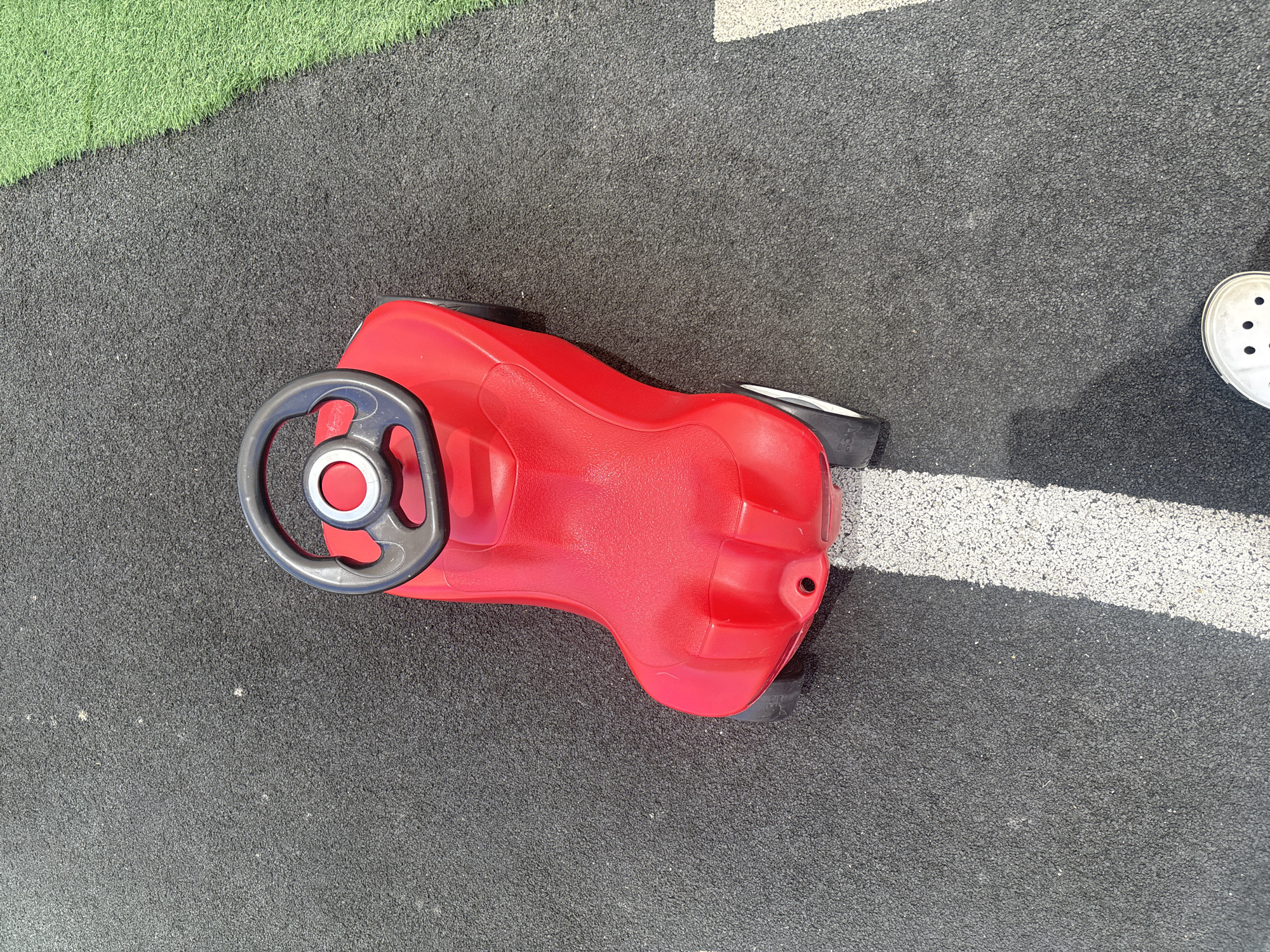 An overhead shot shows a bright red ride-on toy car with a black steering wheel placed on a dark gray, rubberized surface, possibly a playground or park. The car is positioned at an angle, with its front facing towards the lower right of the frame. A white line, characteristic of a road marking, runs diagonally across the dark surface from the bottom left towards the top right. To the right of the dark surface, a strip of vibrant green artificial turf is visible. In the bottom right corner, the toe of a white Croc shoe is partially visible, indicating a person is present. The lighting suggests it is daytime, with shadows indicating the sun's position. There is no clear indication of Dubai, United Arab Emirates in the image itself.